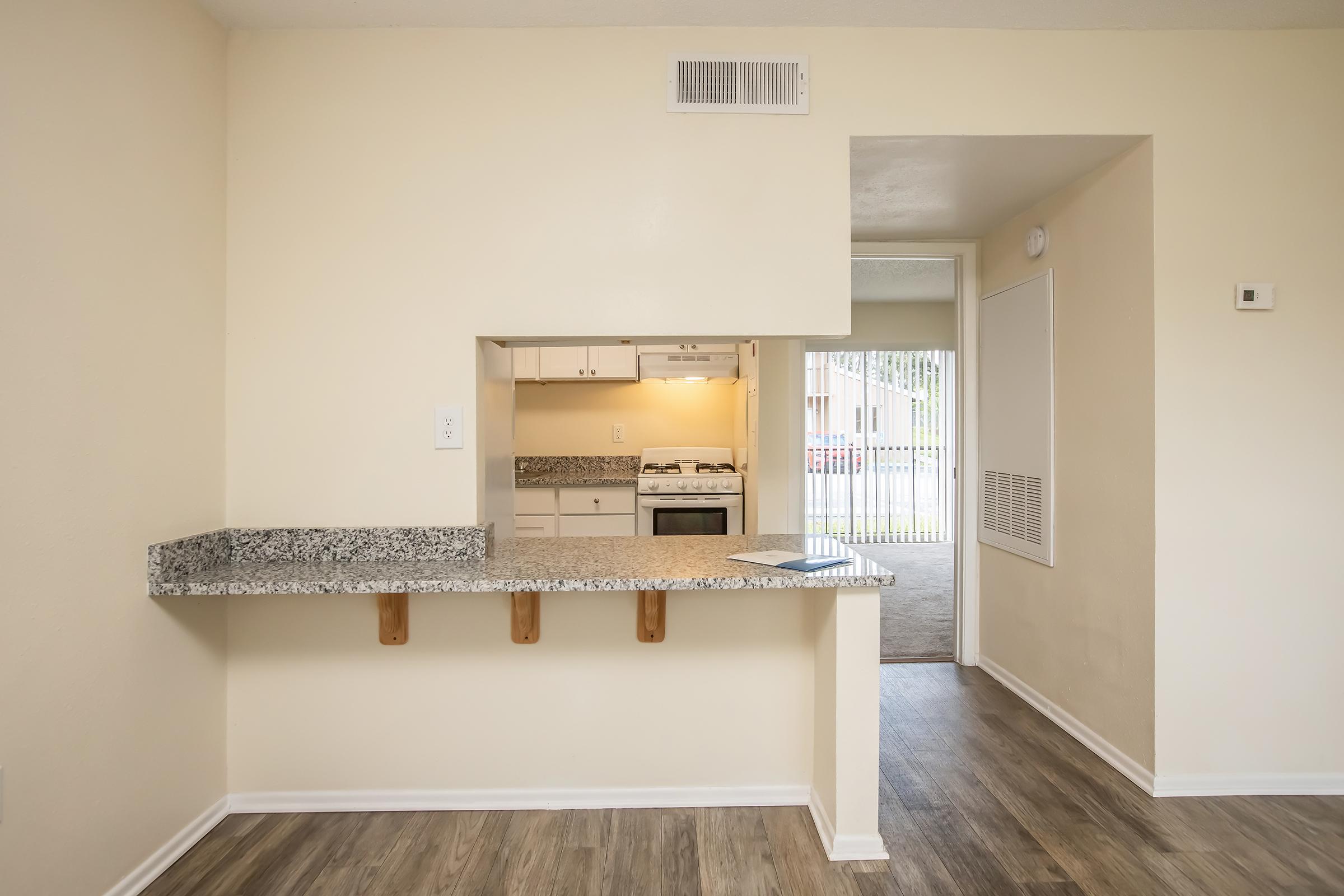 A bright and inviting interior view of a kitchen and living area. The kitchen features a granite countertop with seating, white cabinetry, and a gas stove visible through an open wall cutout. The living area has light-colored walls and wood-like flooring, with a doorway leading to an outdoor area.