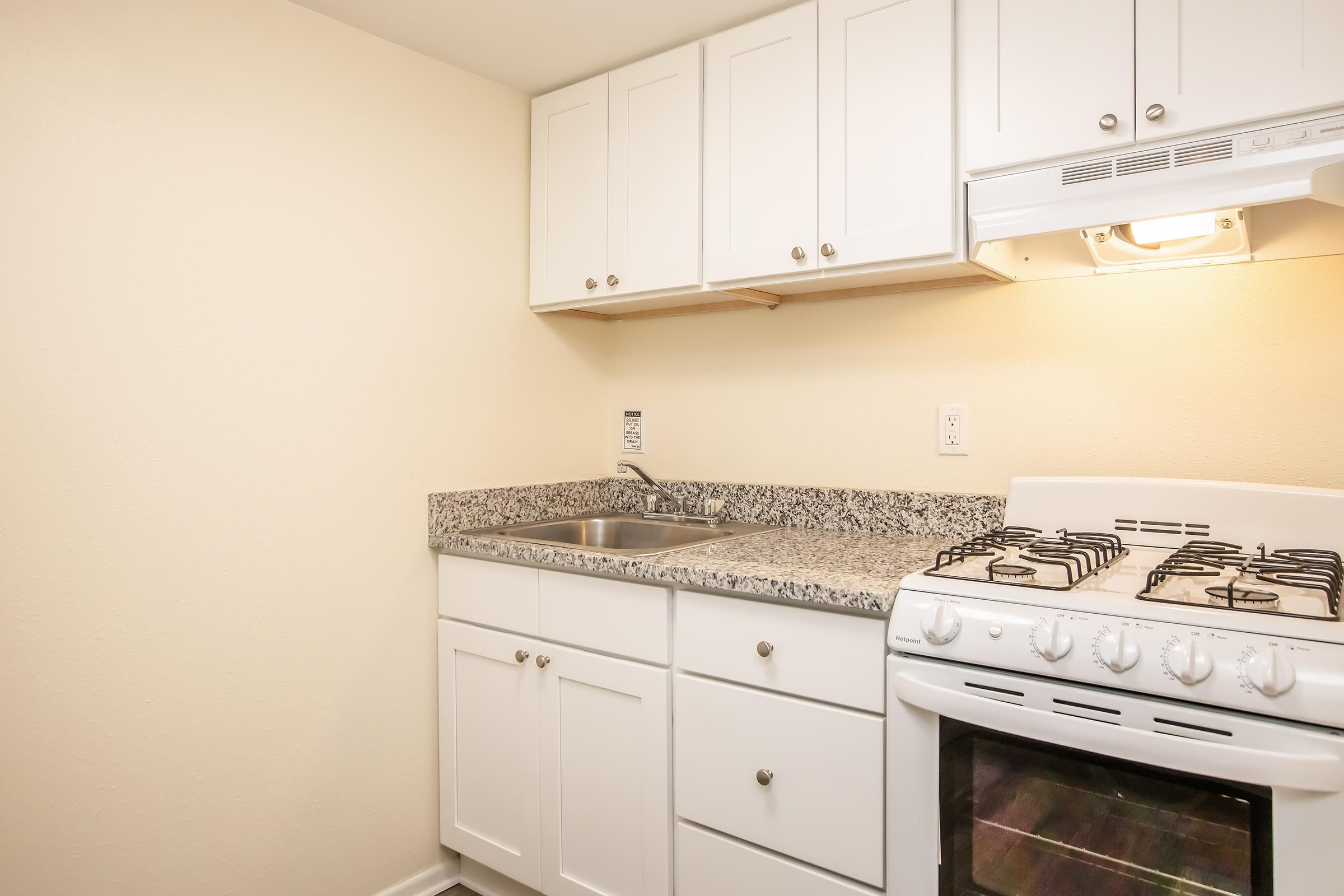 A small kitchen featuring white cabinets, a granite countertop, a stainless steel sink, and a gas stove with an oven. The walls are painted a light color, creating a bright and clean space. The kitchen is functional and compact, suitable for basic cooking needs.