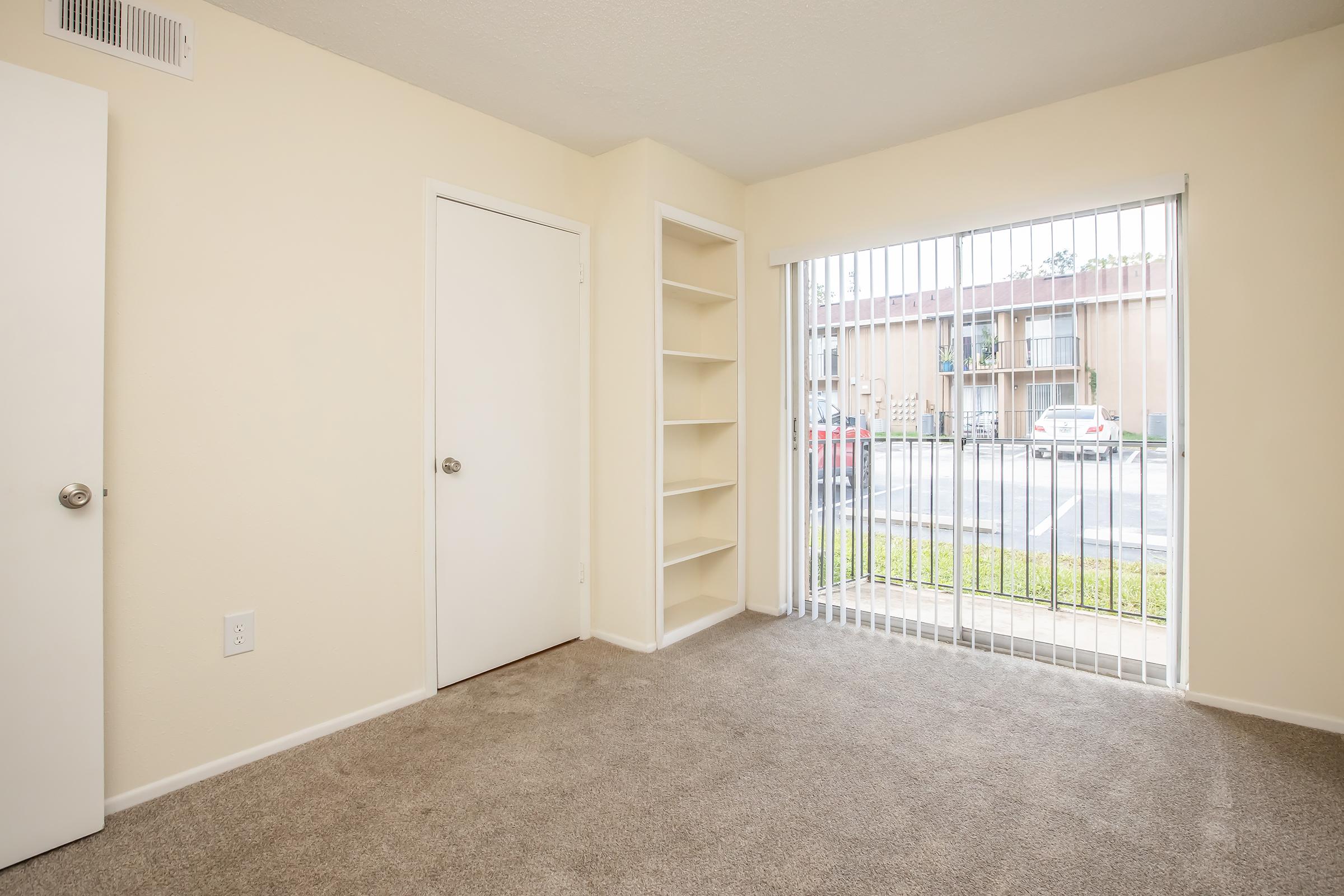 A small room with beige walls and carpet. It has a door on the left and a built-in bookshelf on the right. A large window with vertical blinds offers a view of parked cars outside. Natural light brightens the space, making it feel open and inviting.