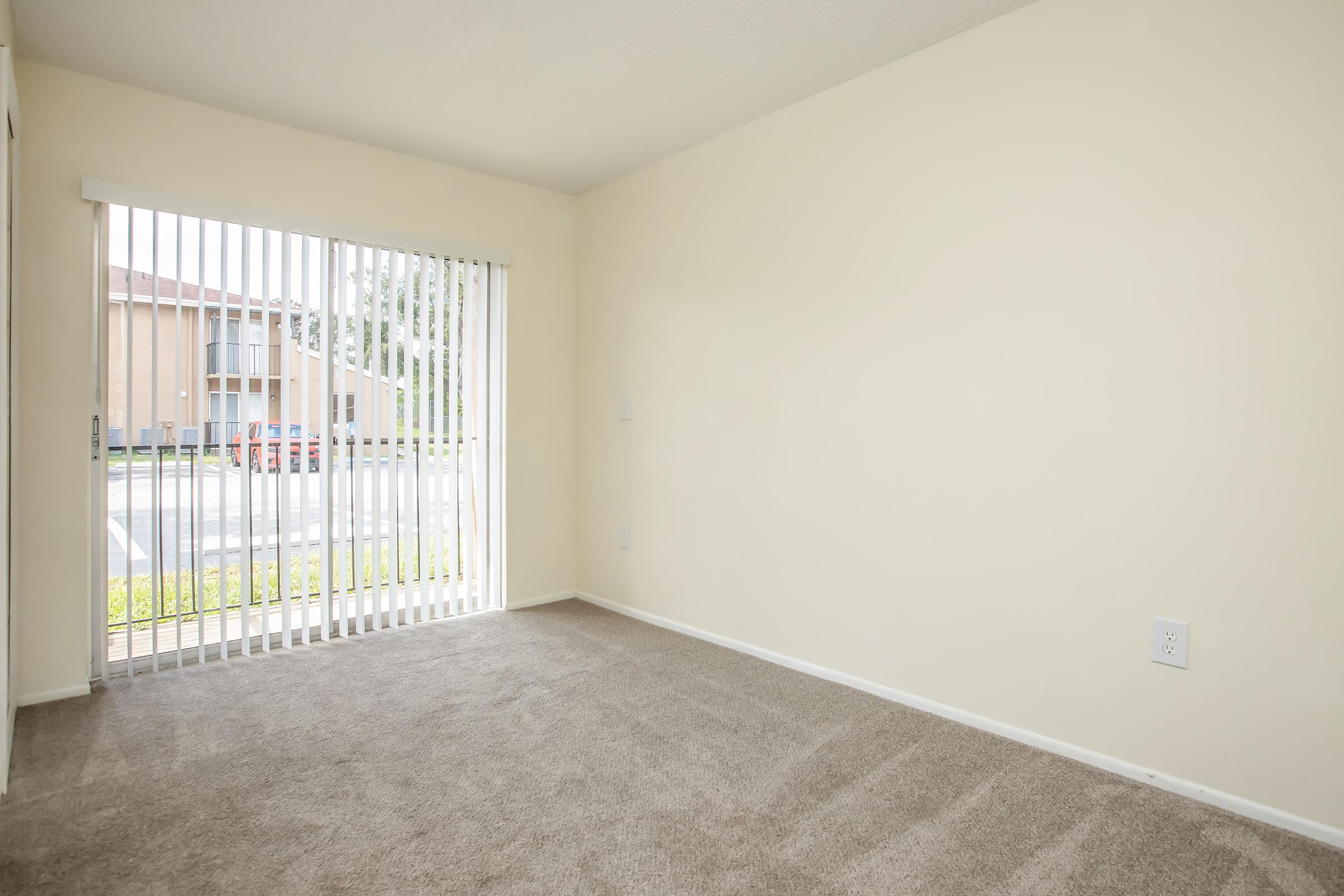 Interior of a vacant room with beige walls and light brown carpet. A large window with vertical blinds allows natural light to enter, revealing an outdoor view. The space appears clean and well-maintained, suggesting it could be used as a living room or bedroom.