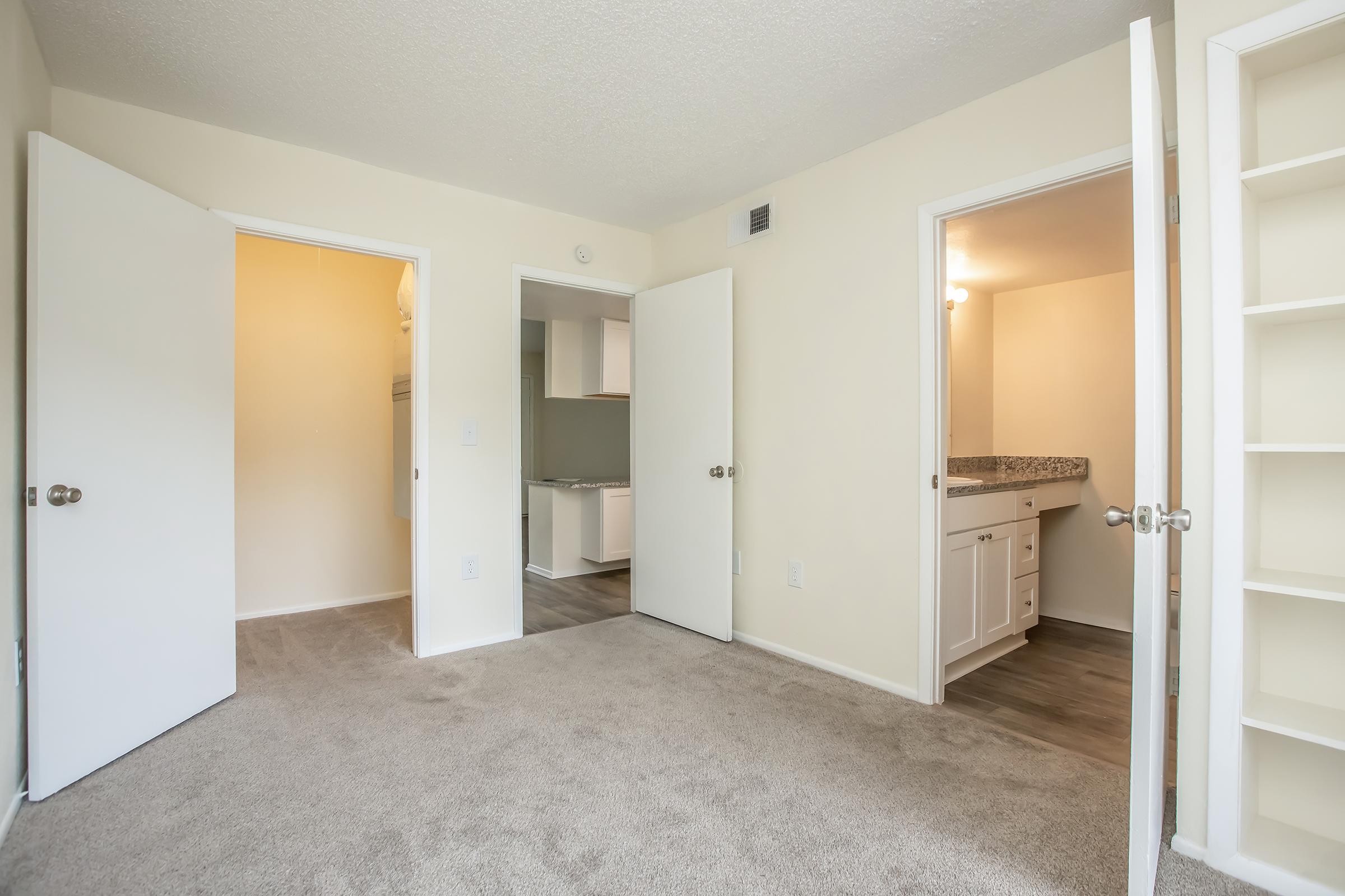 A light-colored room with beige carpet and painted walls. Two white doors are slightly open, leading to adjoining spaces. One door opens to a small bathroom with a sink and a countertop, while the other door leads to a closet or storage area. The overall atmosphere is bright and inviting.