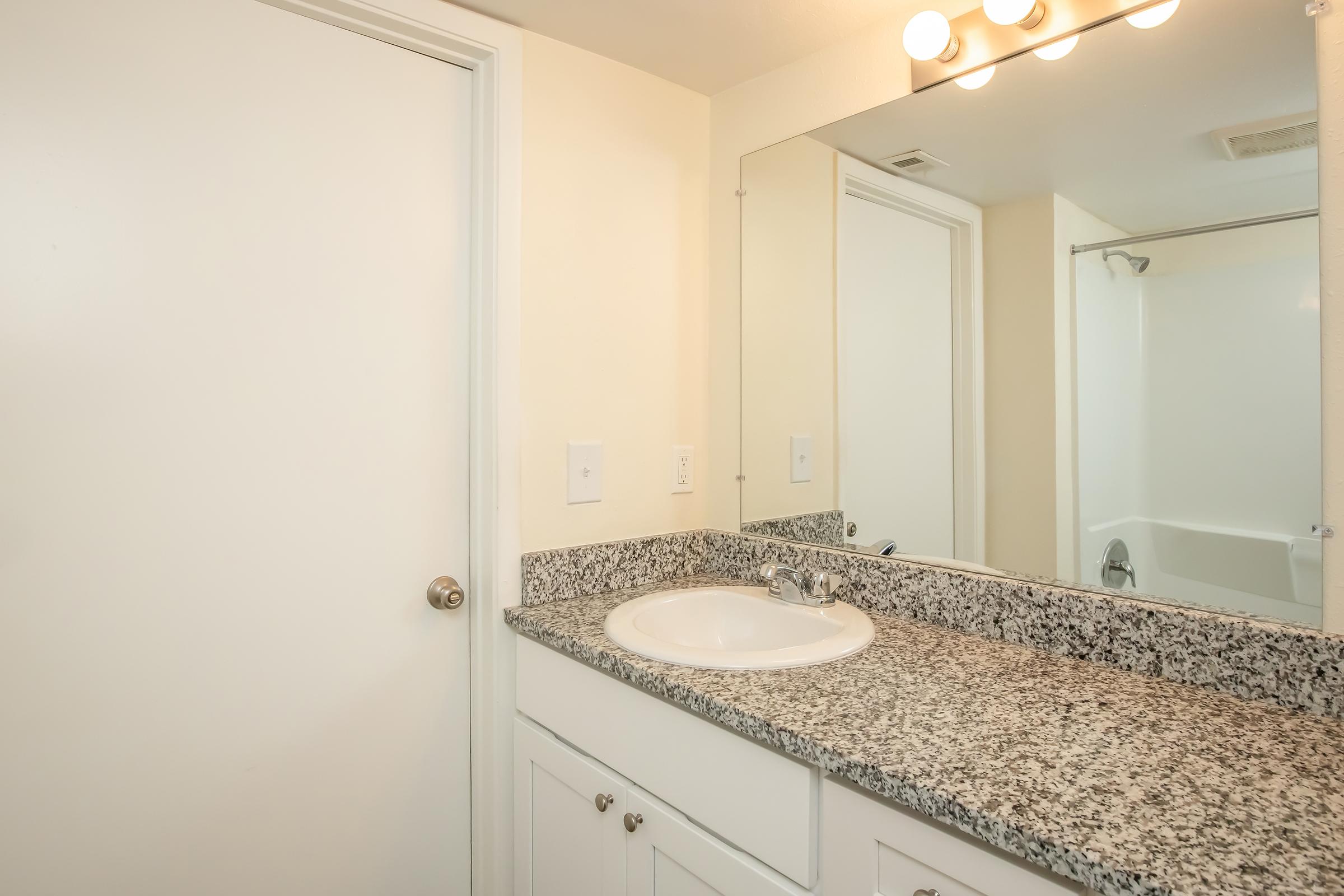 A clean, modern bathroom featuring a granite countertop with a sink, a large mirror above, a light fixture with multiple bulbs, and a beige wall color. A closed white door is visible, along with a bathtub and shower area in the background.