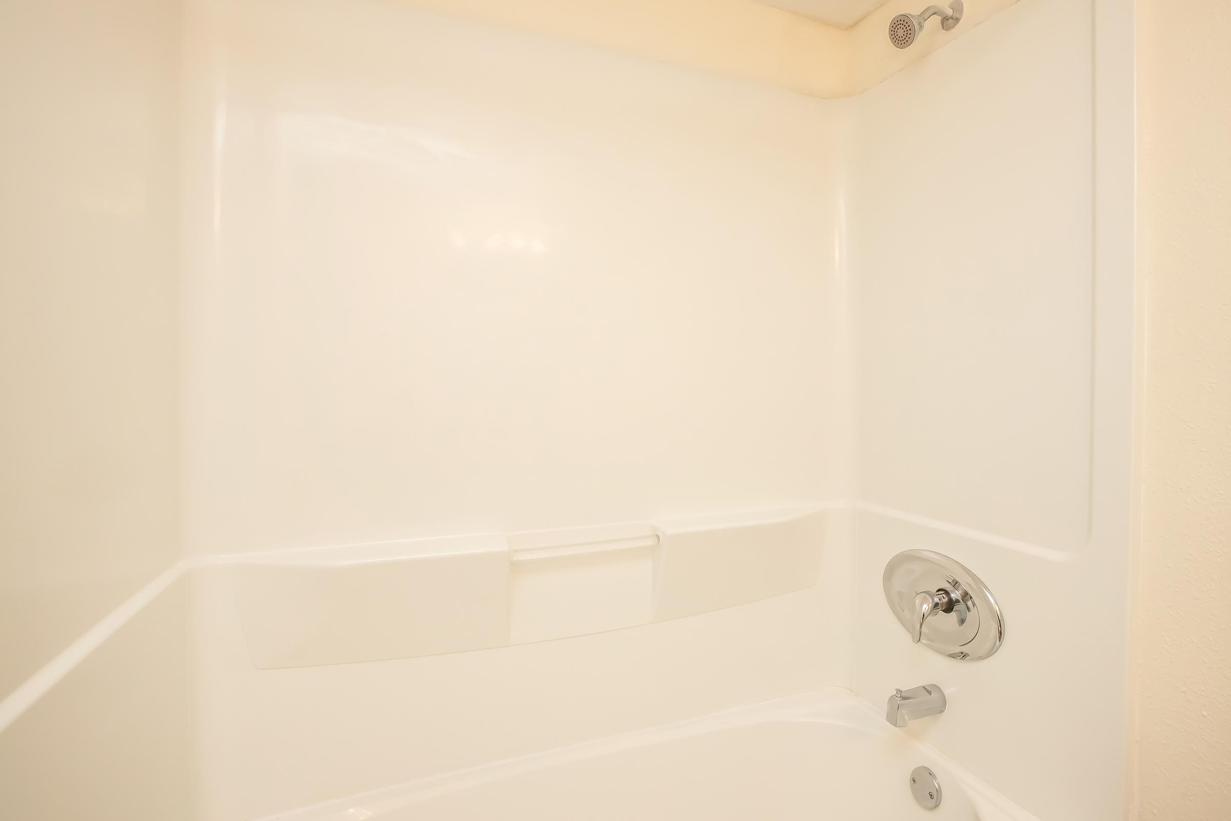 A clean and empty white bathtub and shower enclosure, featuring a smooth wall surface, a showerhead, and a simple chrome faucet. The overall design is minimalist and bright.