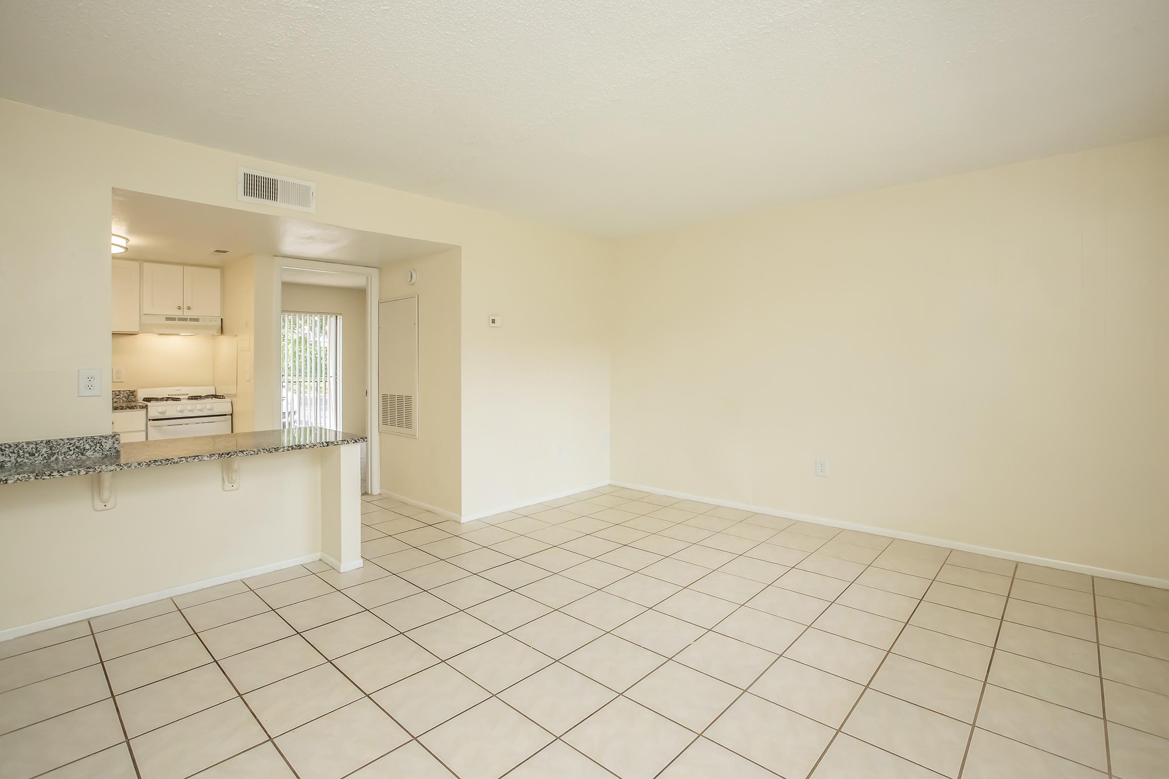 A spacious, empty living area with beige walls and tiled flooring. A kitchen is visible in the background, featuring white cabinets and a granite countertop. The room has a large window with natural light flowing in, creating a bright and airy atmosphere.