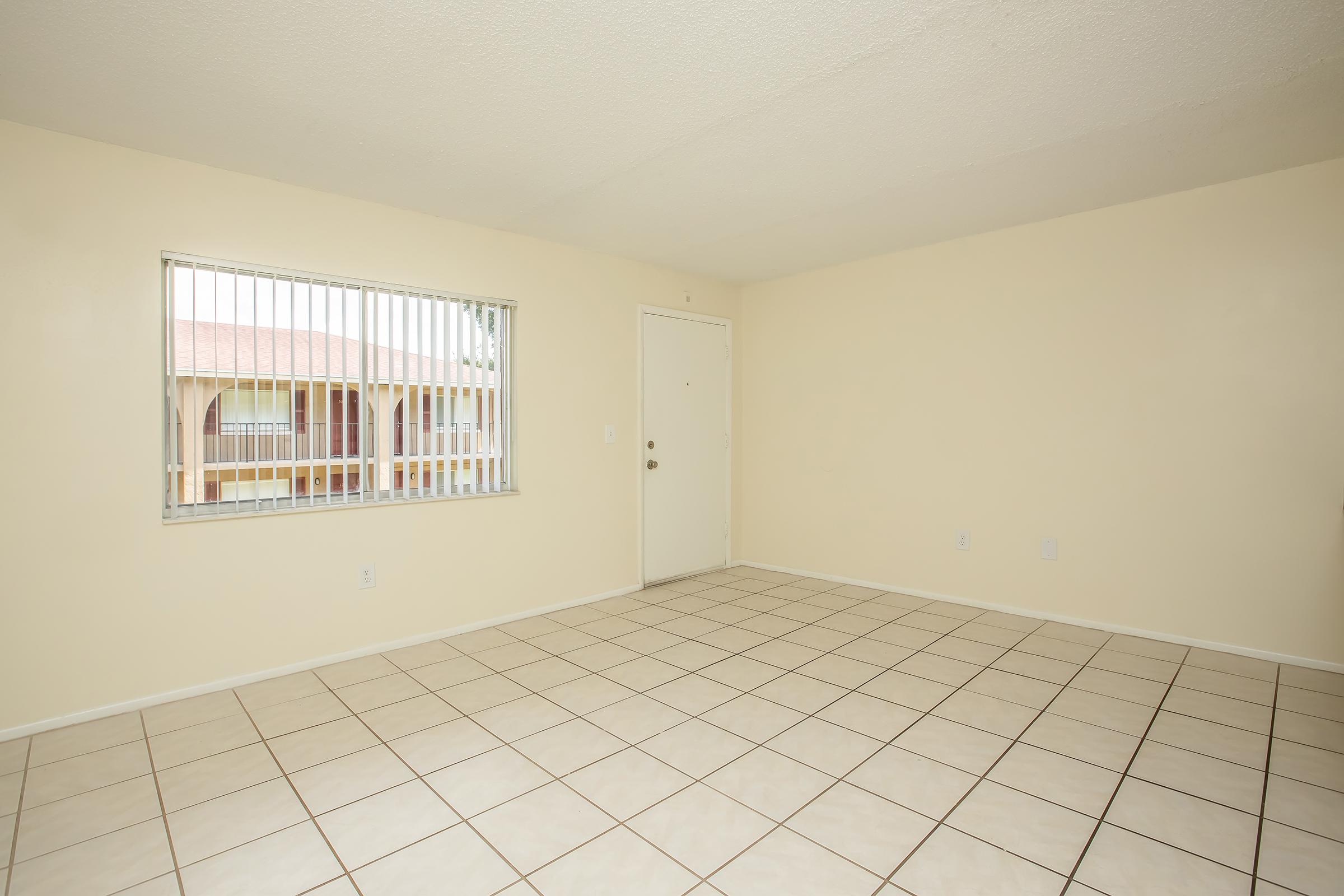Empty room with beige walls and tiled flooring. A window with vertical blinds is on the left, showing a view of an exterior area. A plain door is on the right, leading to another space. The room is well-lit with natural light, creating a spacious and clean atmosphere.