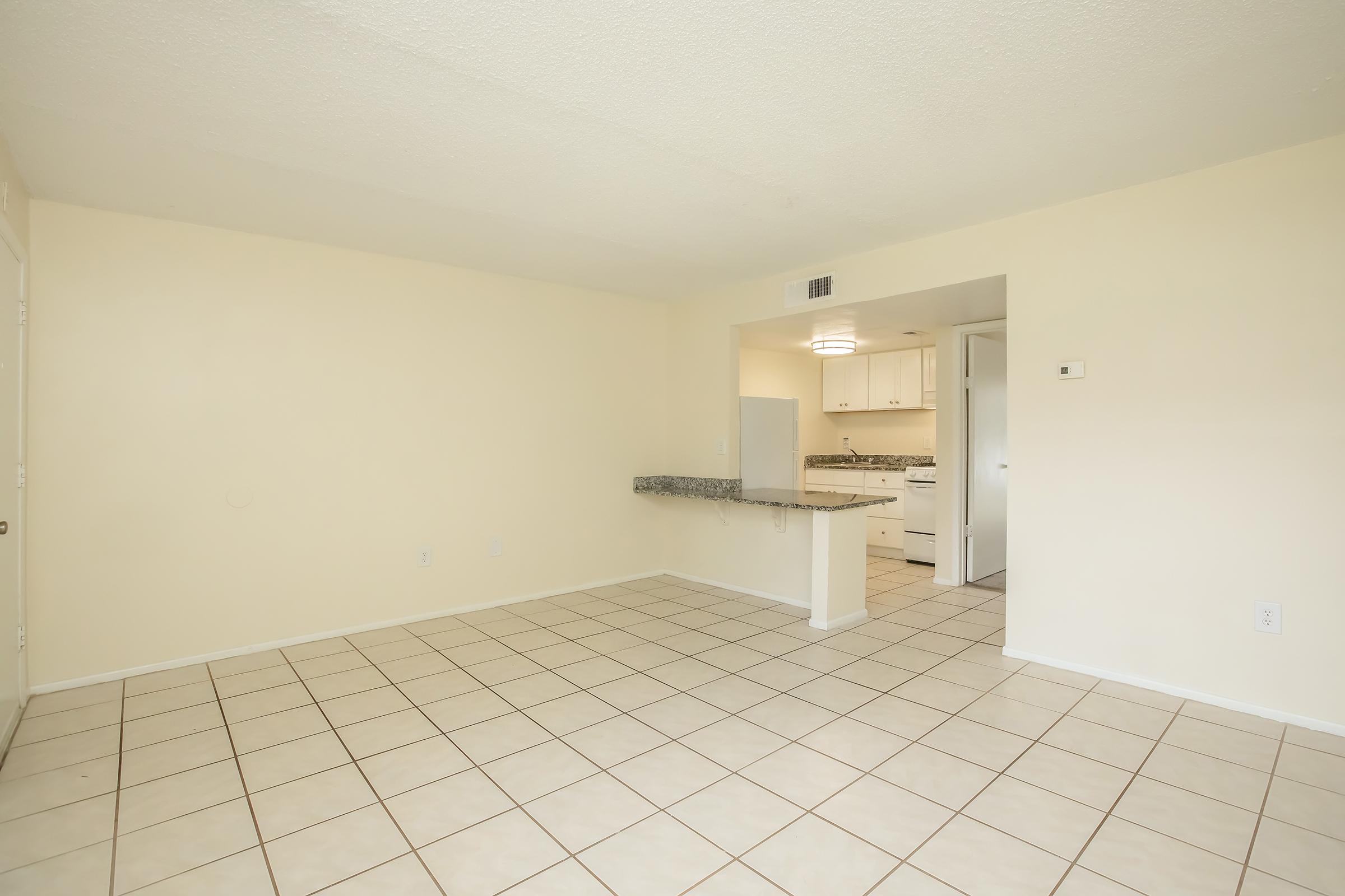 An empty room with light beige walls and tiled flooring. To the left, a door leads outside. In the background, there is a kitchen area with white cabinets and a countertop. Natural light brightens the space, creating an inviting atmosphere.