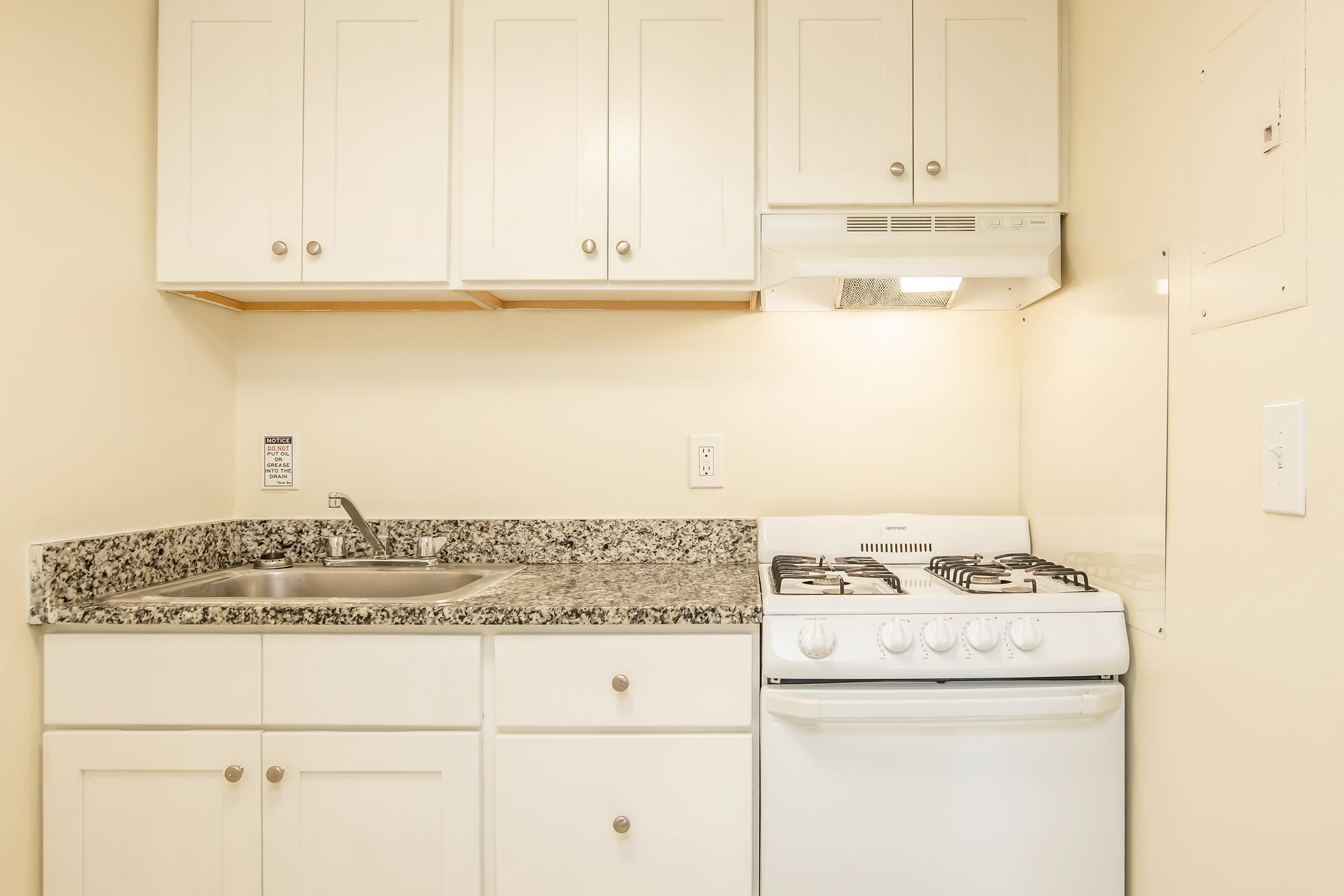 A small kitchen featuring white cabinets, a stainless steel sink, and a gas stove with a hood. The countertops are granite, and the walls are painted a light color, creating a bright and clean appearance. There's an electrical panel visible on the right side of the image.