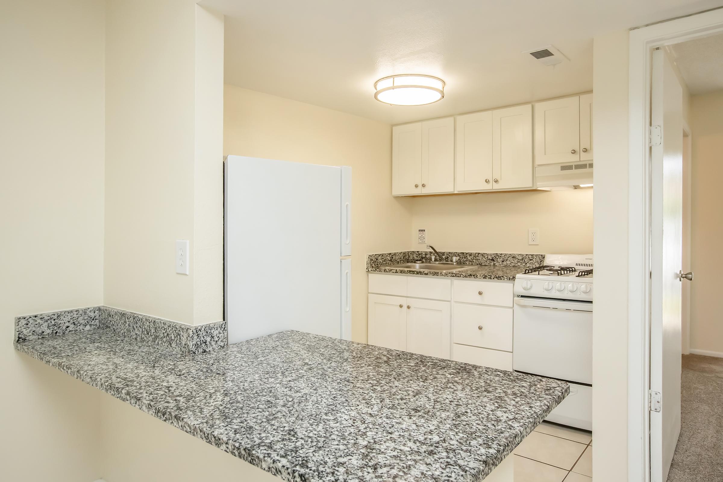 A modern kitchen featuring white cabinetry, a granite countertop, and stainless steel appliances. The space includes a stove, refrigerator, and ample storage. Natural light from a ceiling fixture illuminates the area, creating a bright and inviting atmosphere. A bar area connects to another room.