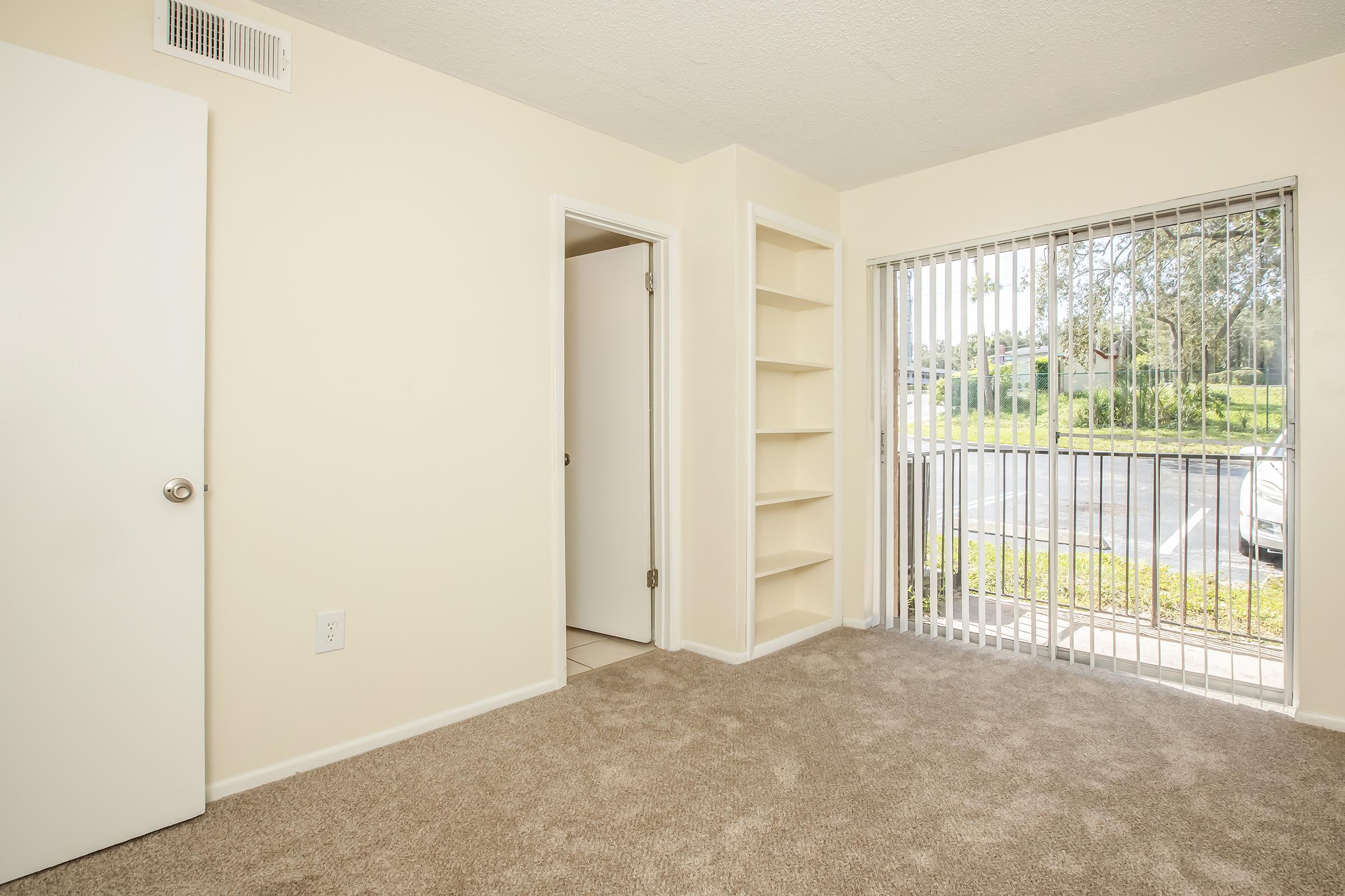 Bright, empty room with beige walls and plush carpet. A white door leads to a closet, and another door opens to the hallway. There's a built-in bookshelf along one wall, and large windows with vertical blinds allow natural light to fill the space. Overlooks a green area with trees outside.