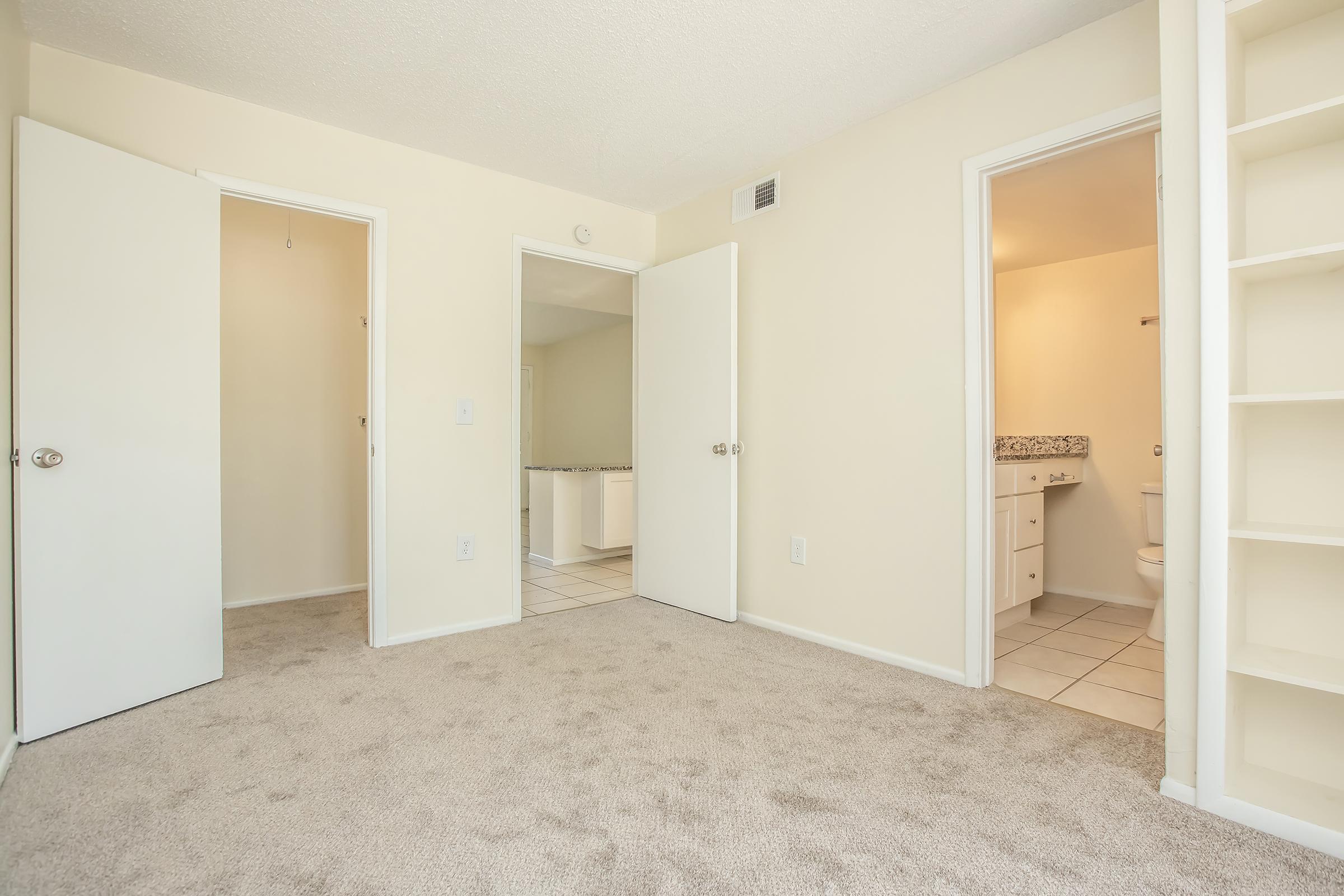A spacious room with beige walls and carpet, featuring two open doors leading to adjoining rooms. One door reveals a bathroom area with tiled flooring, while the other leads to another room. Natural light brightens the space, which also includes a built-in shelf along the wall.