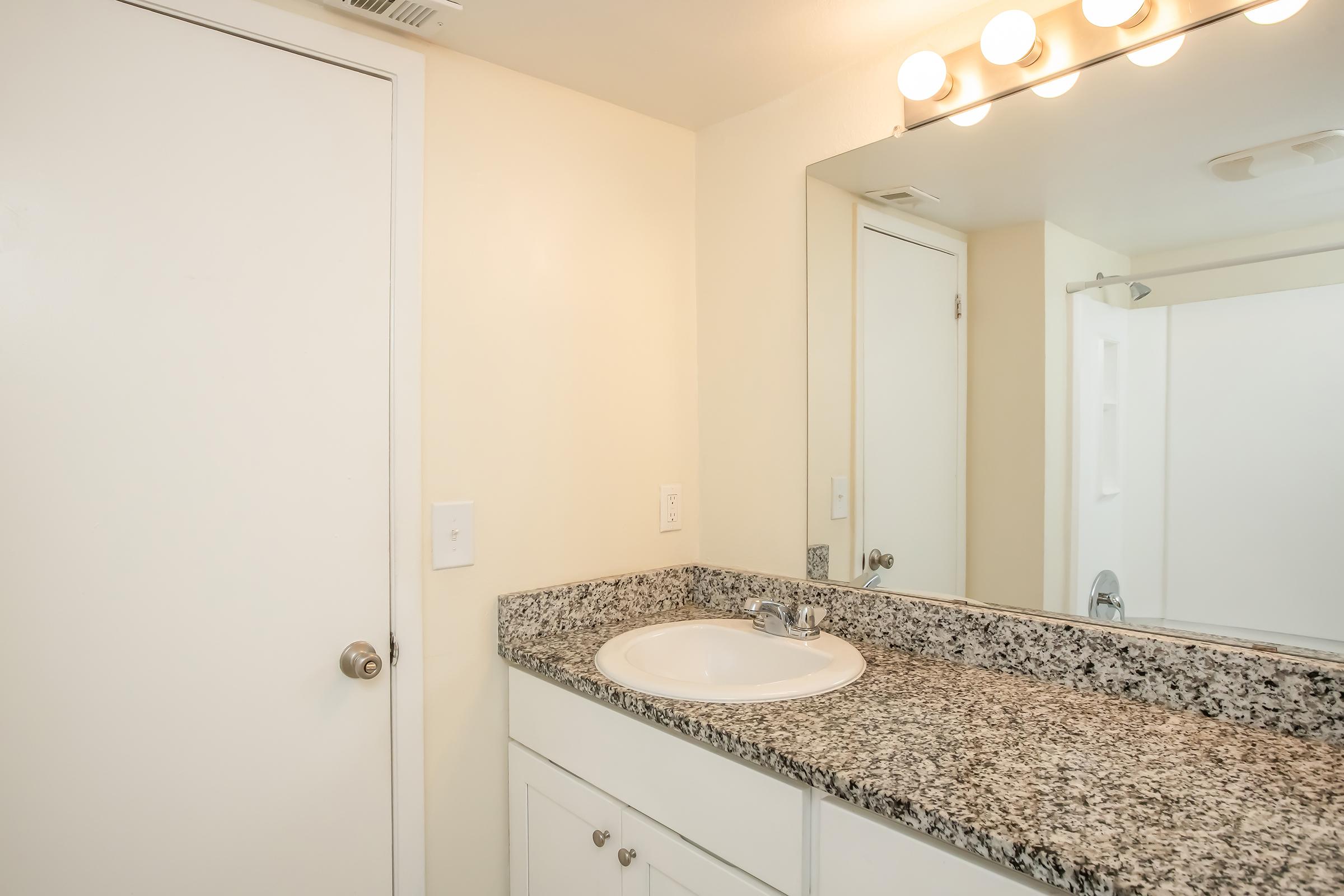 A clean, modern bathroom featuring a light-colored wall with a round mirror above a granite countertop. The countertop has a white sink and faucet, while a closed door is visible on the left. A shower area is partially visible in the background, highlighting a spacious and uncluttered environment.