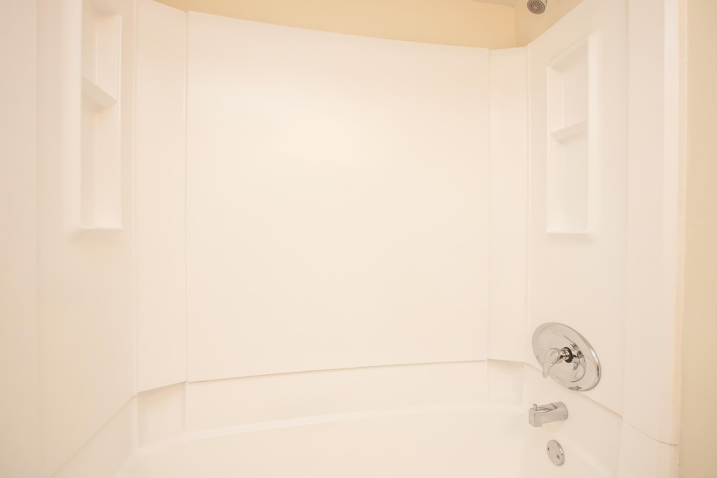 Interior view of a clean, modern shower stall with smooth white walls and built-in shelves. The faucet and showerhead are visible, featuring a chrome finish. The overall design is simple and minimalistic, emphasizing a bright and spacious feel.