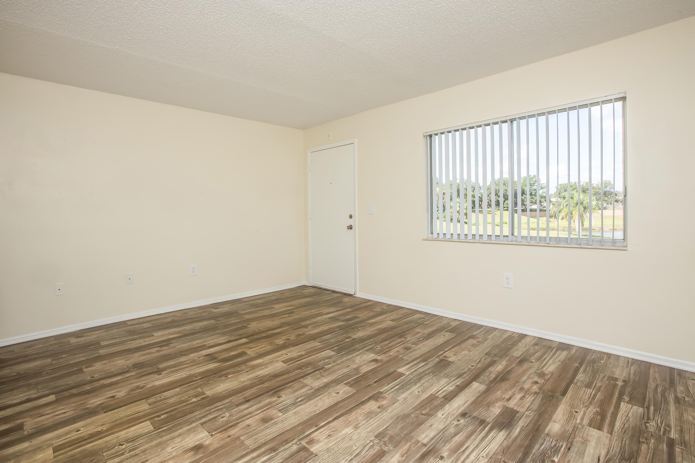 A spacious, empty room with light beige walls and a large window featuring vertical blinds. The floor is covered with wooden laminate, giving a warm appearance. There is a door on one wall, and the overall ambiance is bright and inviting.