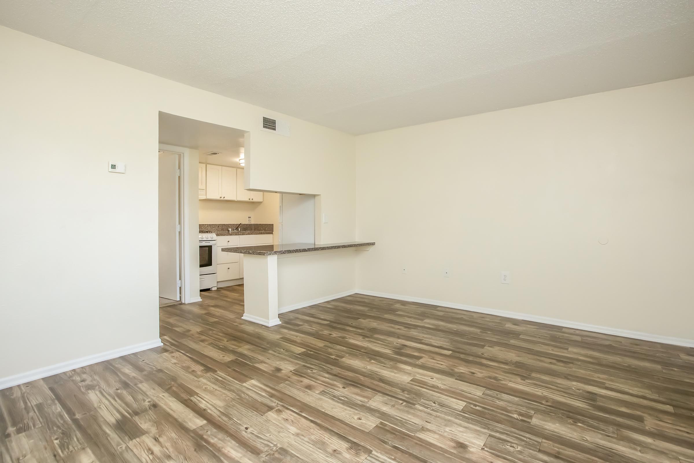 Bare interior of an empty room featuring light-colored walls and a wooden floor. A small opening leads to a kitchenette with white cabinets and a stove visible in the background. The space is bright and uncluttered, suitable for various furniture arrangements.