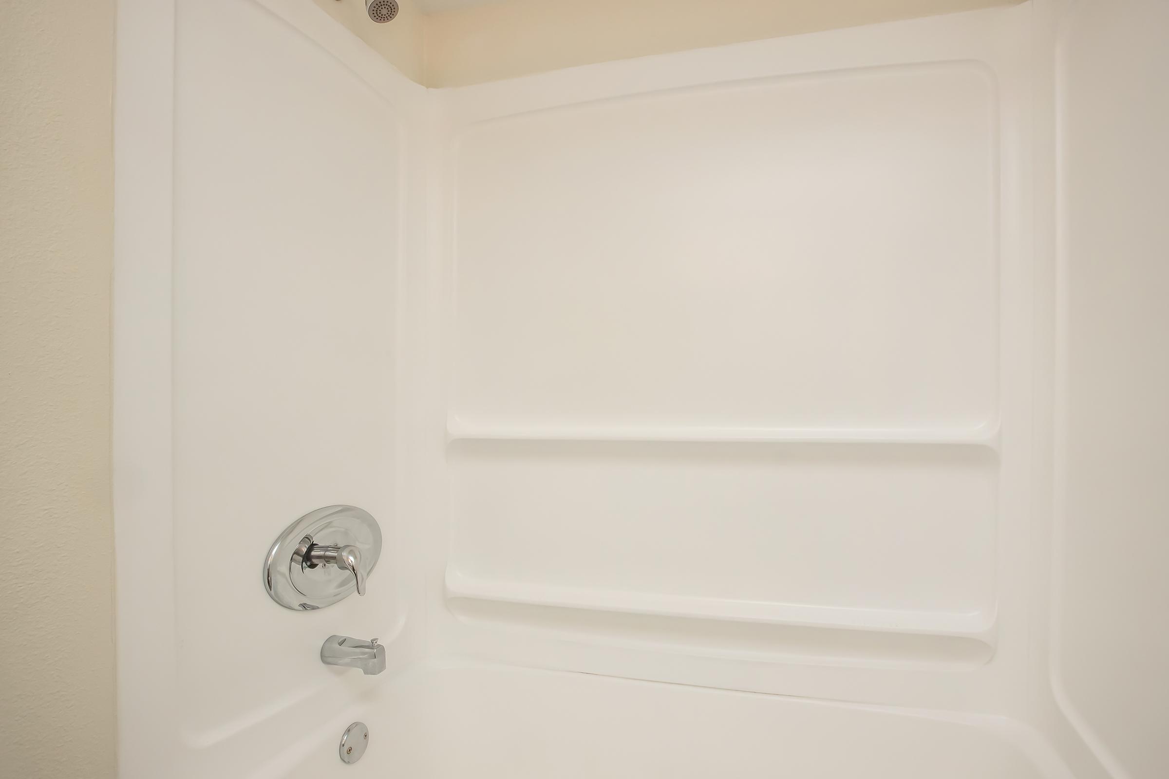 A clean, white shower stall featuring smooth walls, a chrome faucet, and a shower handle. The design is modern and minimalistic, with two built-in shelves for storing toiletries. The ambient lighting enhances the bright appearance of the space.