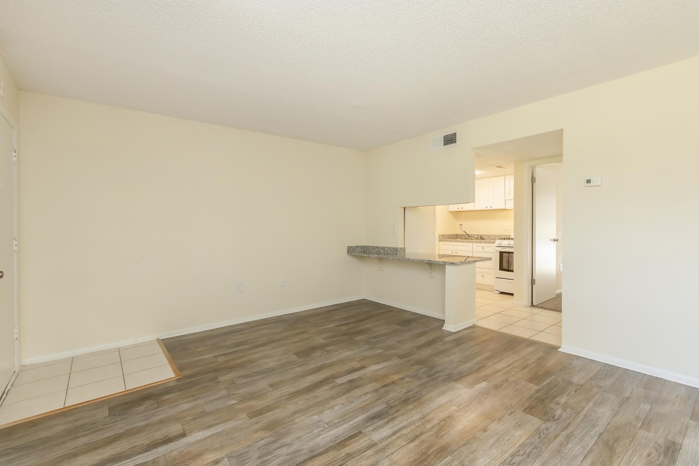 Empty living space with light-colored walls and laminate flooring. A small kitchen area is visible in the background, featuring white cabinets and appliances. The entrance is to the left, with a tiled area. Natural light brightens the room, creating a spacious and airy feel.