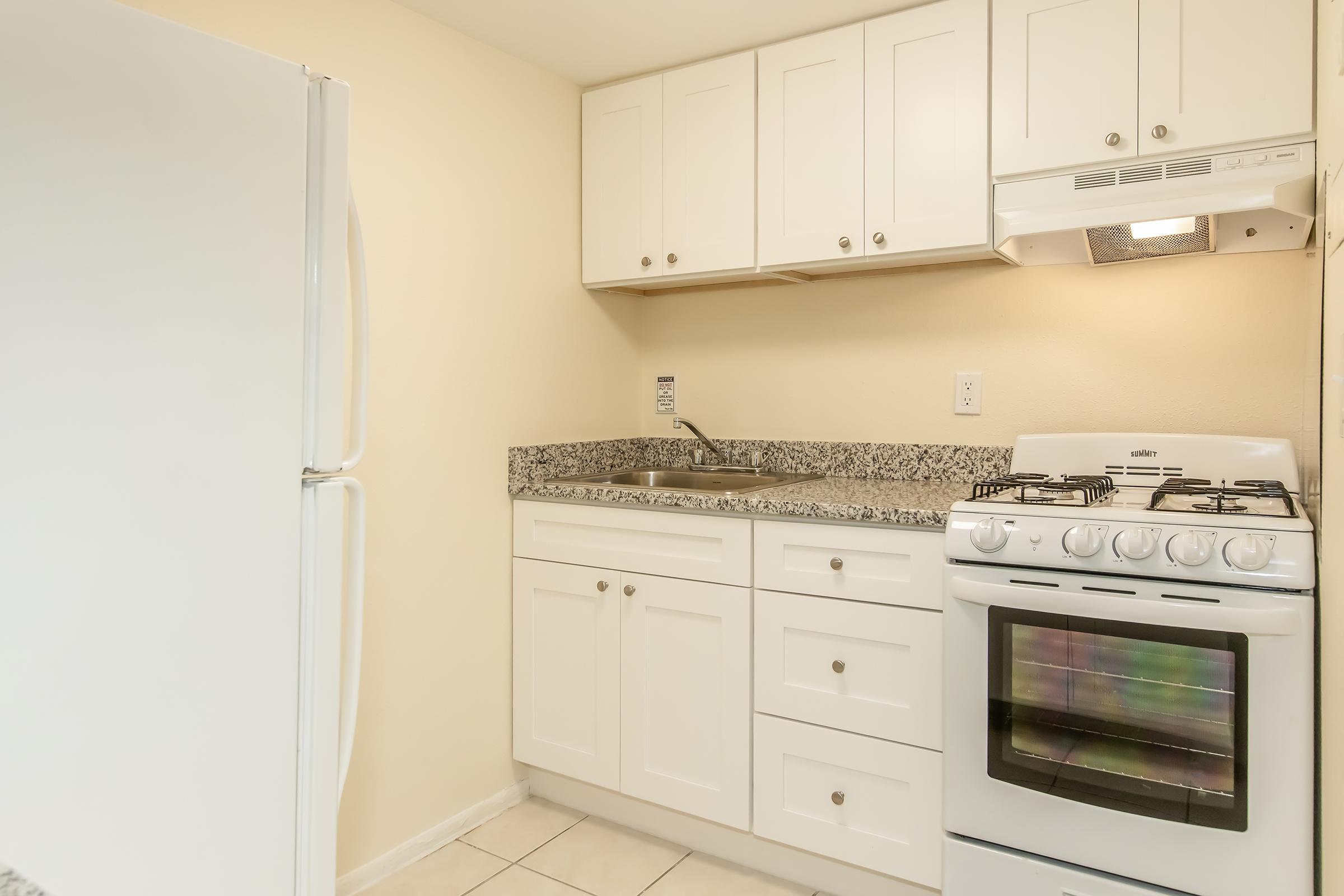 Small kitchen with white cabinetry and granite countertops. Features a stainless steel gas stove with an exhaust hood and a sink. A white refrigerator is on the left, and there are light-colored tiles on the floor. The walls are painted a neutral beige.