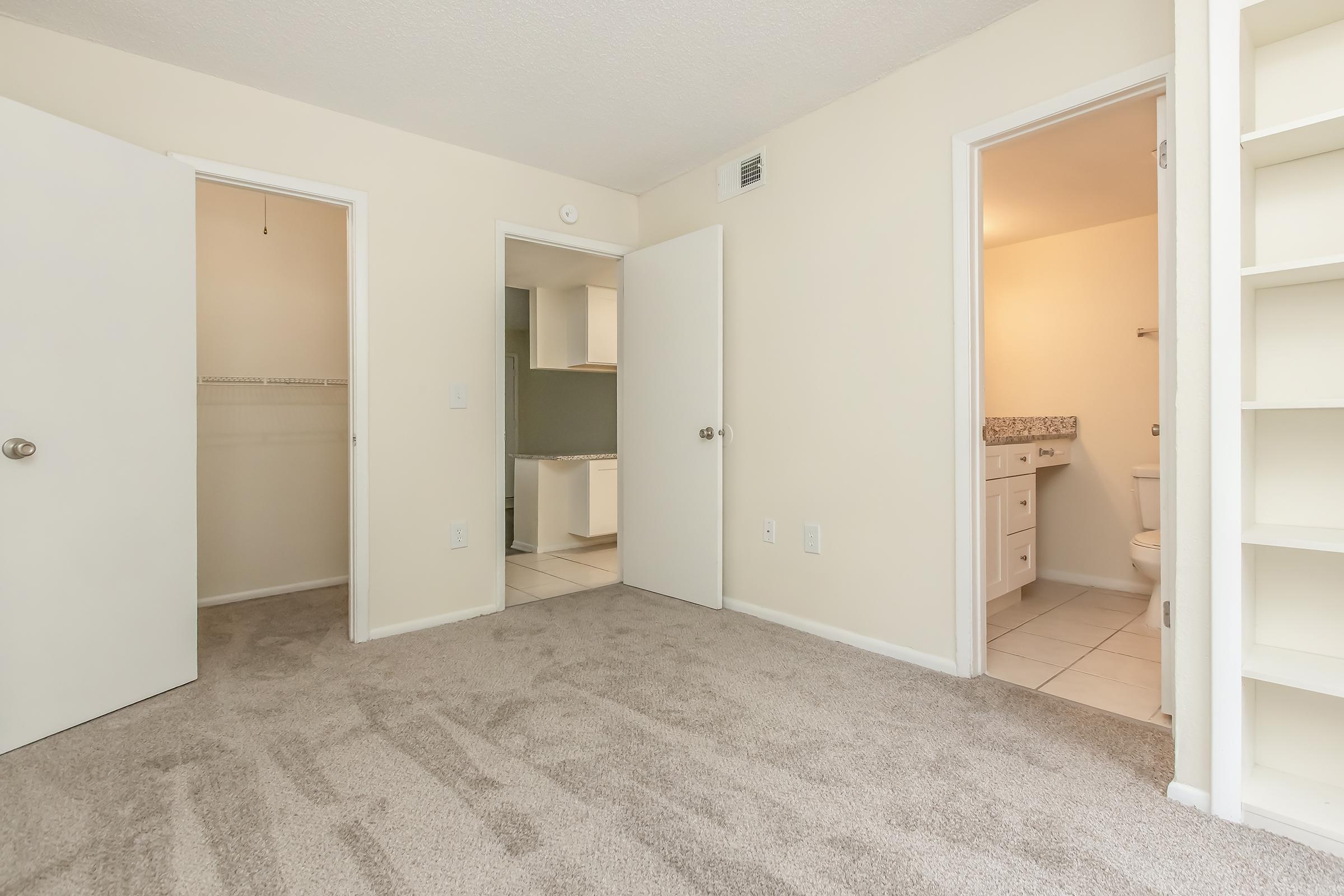 A clean and empty room featuring light-colored walls, soft carpet flooring, and two open doors leading to a closet and bathroom. The room is well-lit and spacious, with additional shelving on one wall.