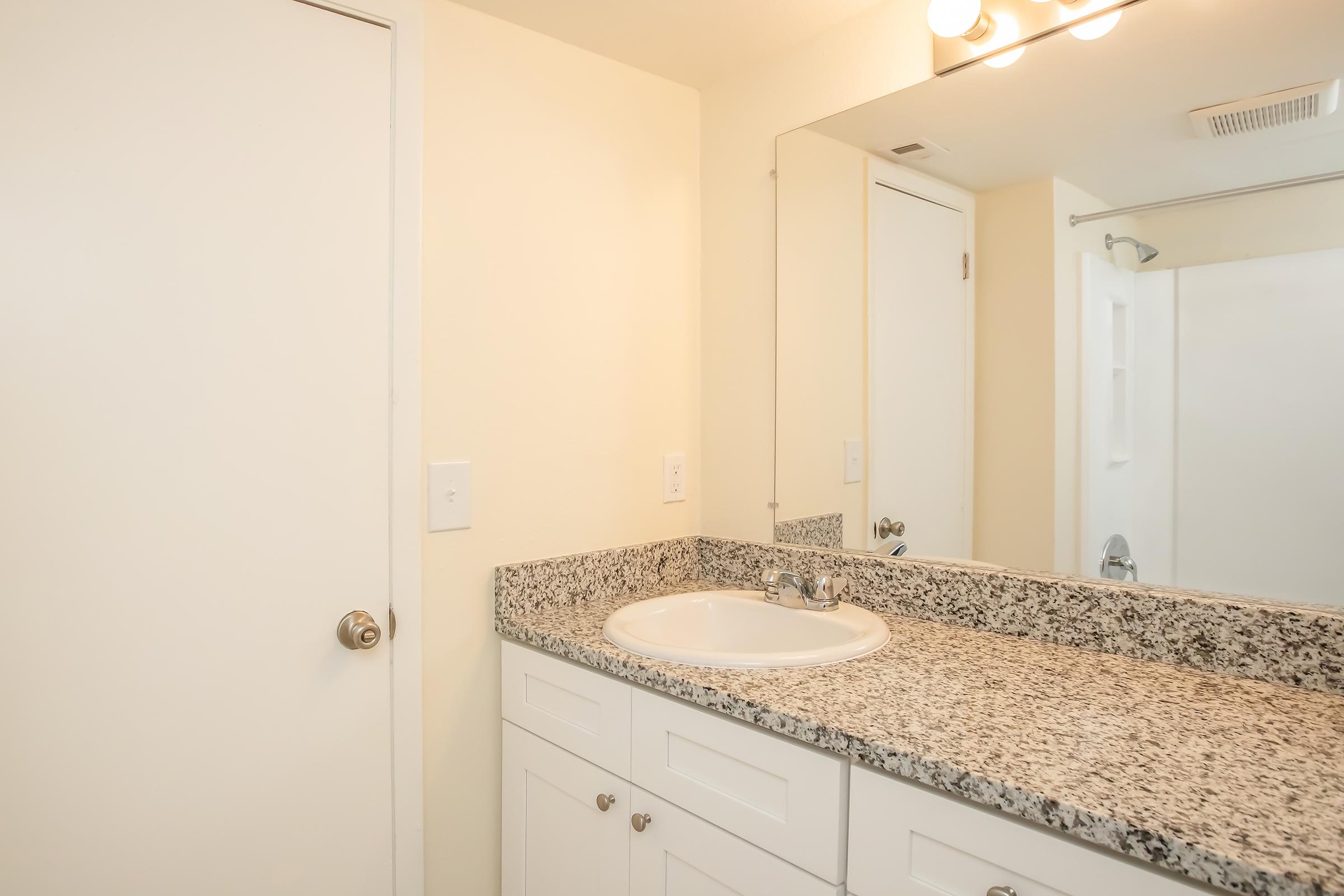 A bathroom featuring a countertop with a sink, a mirror above the sink, and a door leading to another space. The walls are painted a light color, and the countertop has a granite finish. A shower area is visible in the background, with a white curtain or door.
