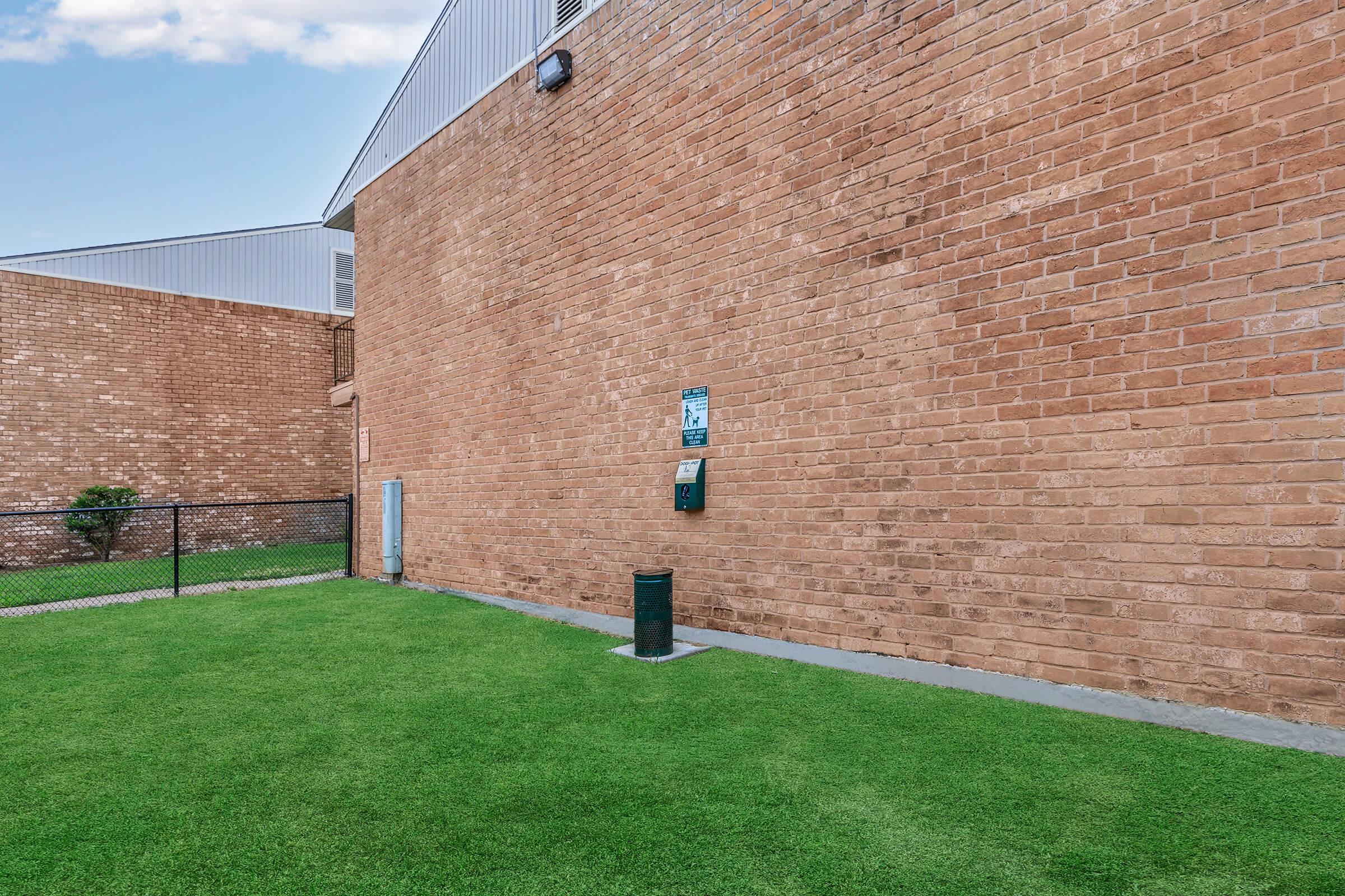 A green grass area beside a brick wall of a building, featuring a water fountain mounted on the wall and a cylindrical trash can on the ground. A fence is visible in the background, and the sky is partly cloudy.