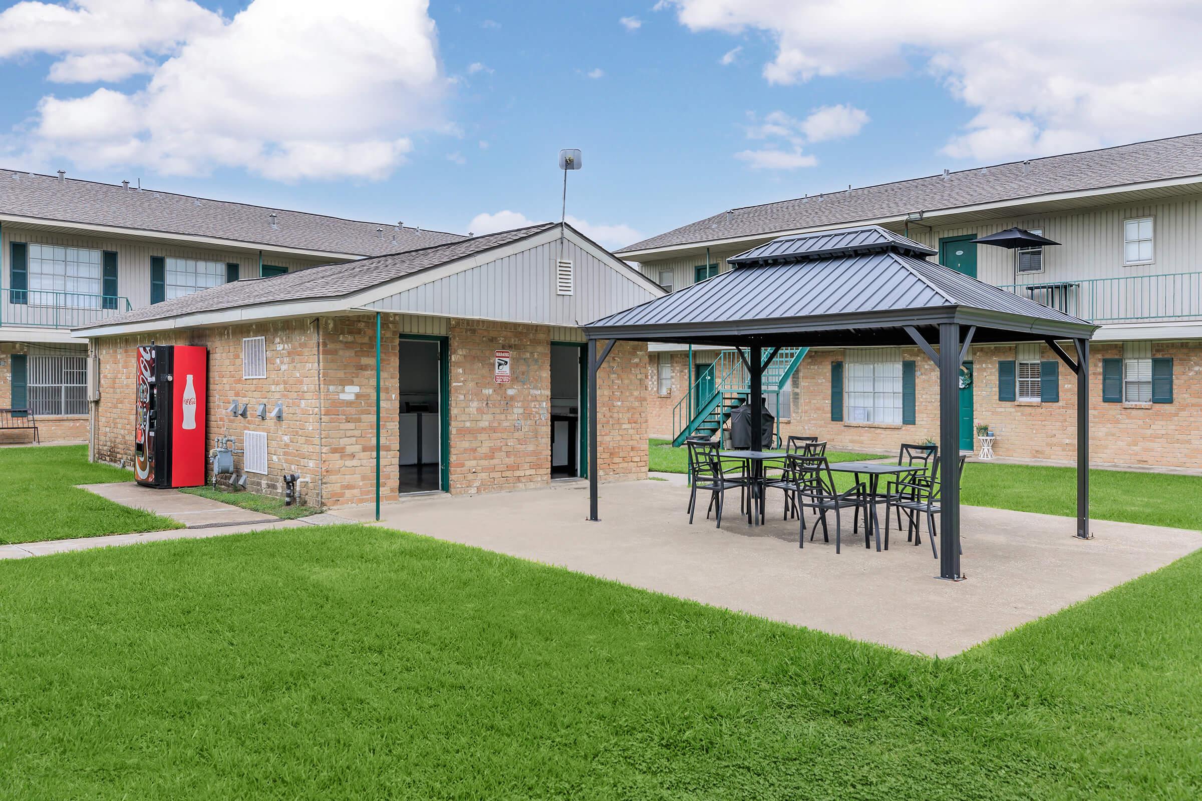 A courtyard area featuring a gazebo with seating and tables, surrounded by green grass. In the background, a brick building is visible, along with a red vending machine. The scene is set in a residential complex with multiple stories and a staircase visible. The sky is partly cloudy.