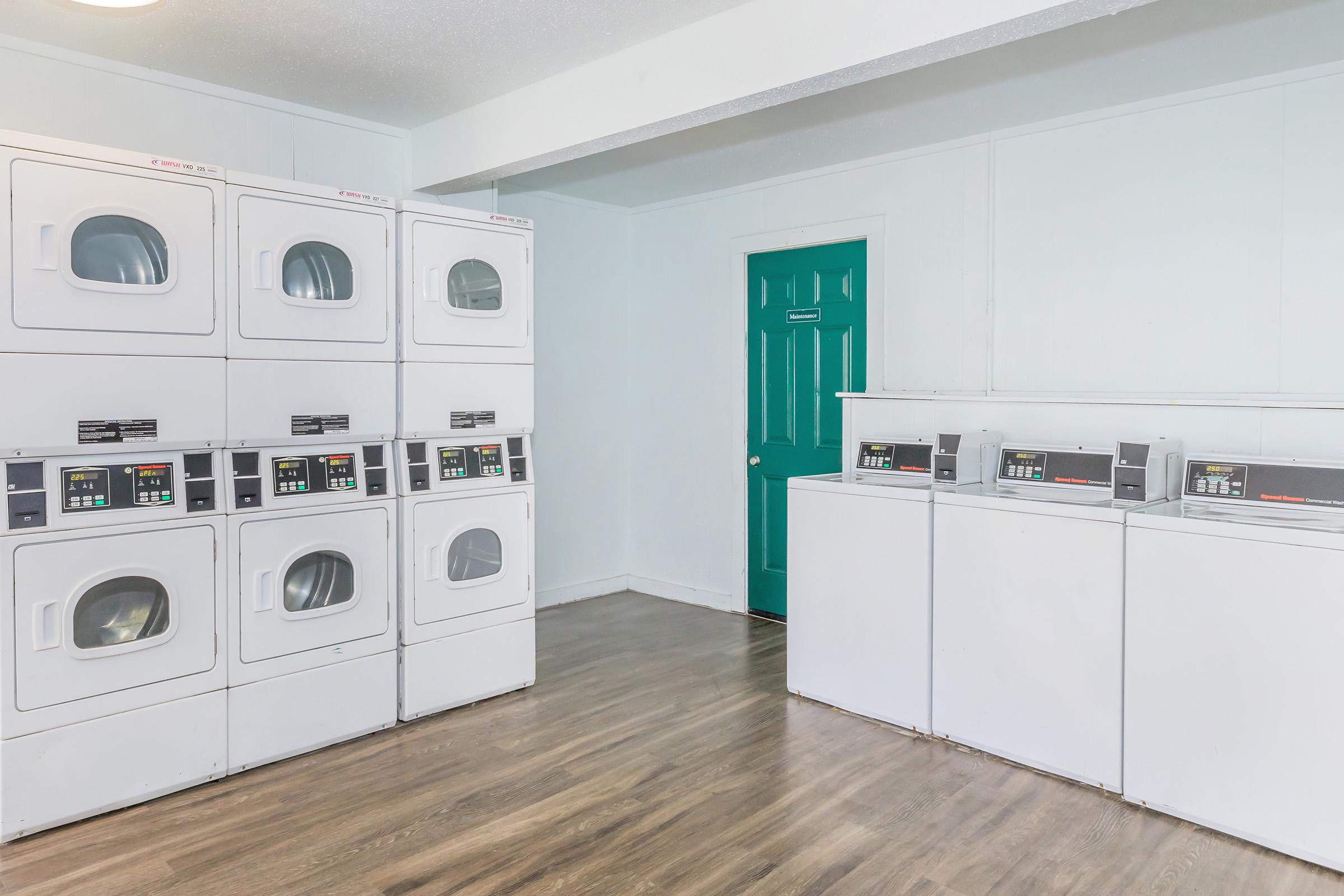 A clean laundry room featuring stacked white dryer units on the left and two front-loading washing machines on the right. The room has light-colored walls and wooden flooring, with a bright teal door at the back. Bright and organized environment, ideal for laundry tasks.