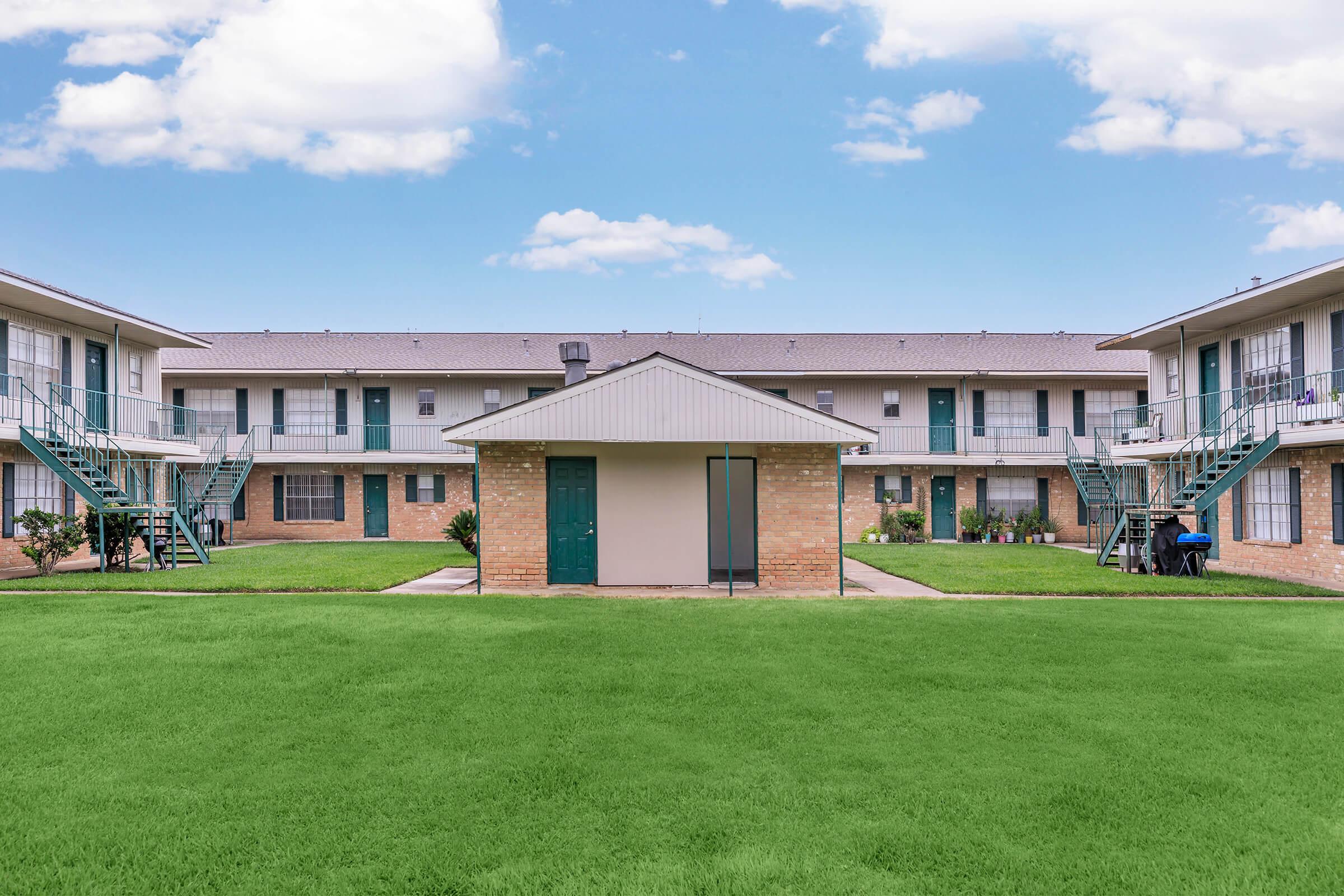 A view of a multi-unit apartment building with a central green lawn. The building has two levels with multiple entrances, surrounded by blue sky and clouds. There are staircases on either side leading to the upper level, and a patio area with seating visible in the background.