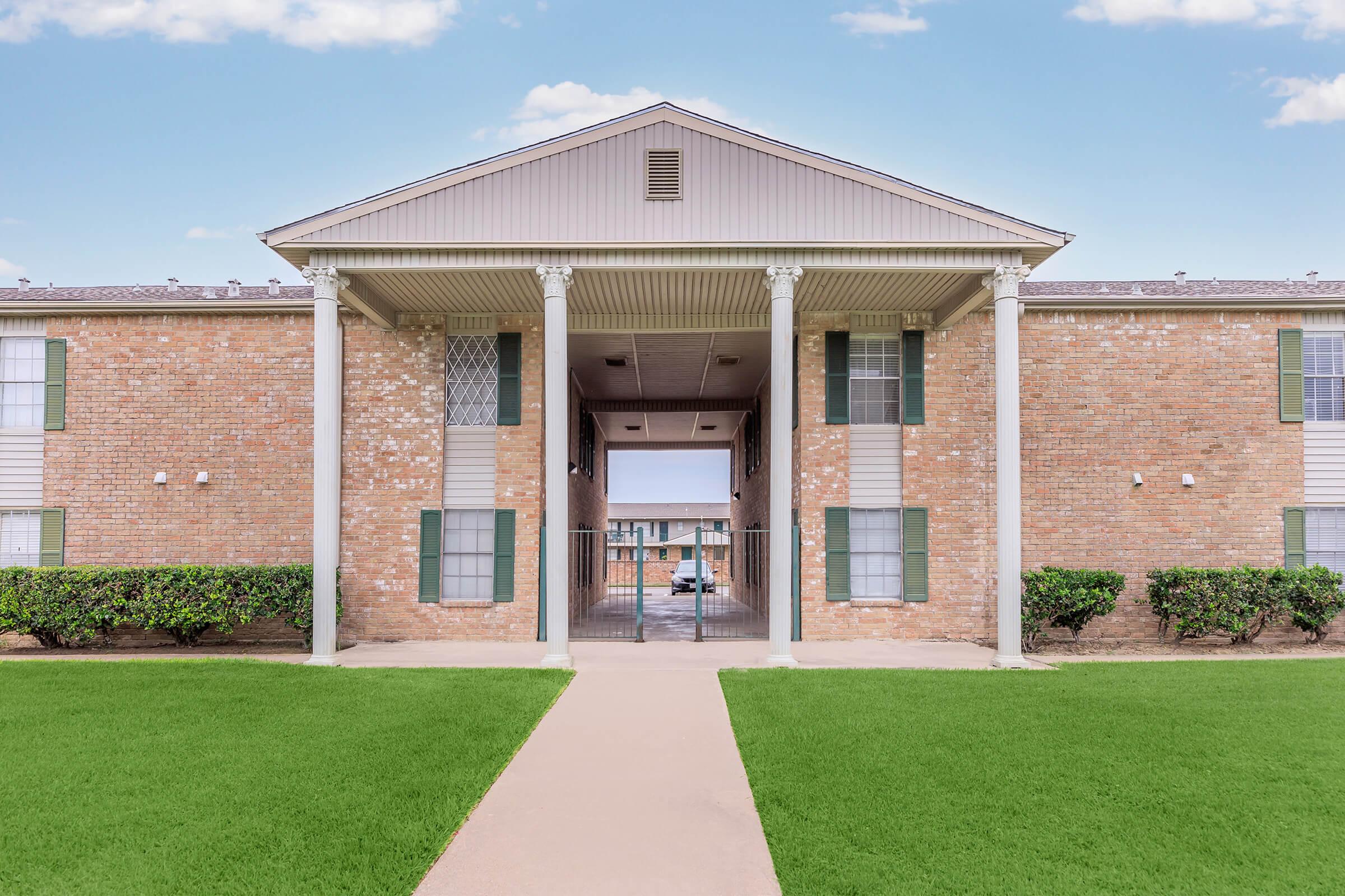 Exterior view of a brick apartment building featuring a central entrance with pillars, flanked by green shrubbery and a pathway. The sky is clear, and there are additional units visible in the background.