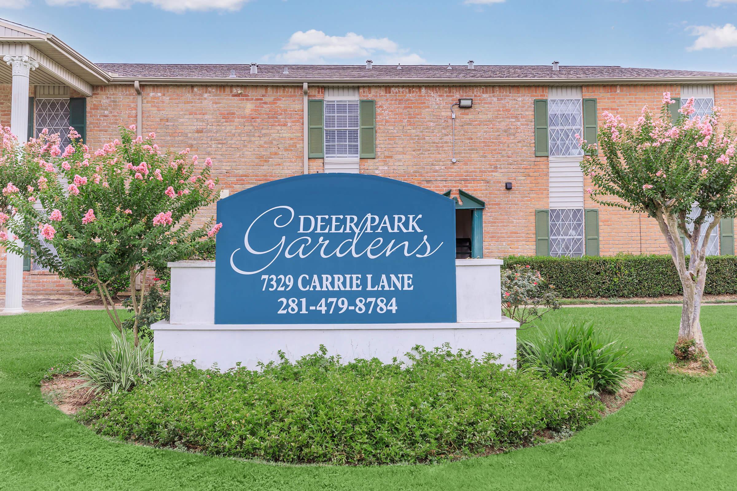 Sign for Deer Park Gardens located at 7329 Carrie Lane, with the contact number 281-479-8784. The sign is surrounded by flowering plants and greenery, with a brick building and blue sky in the background.