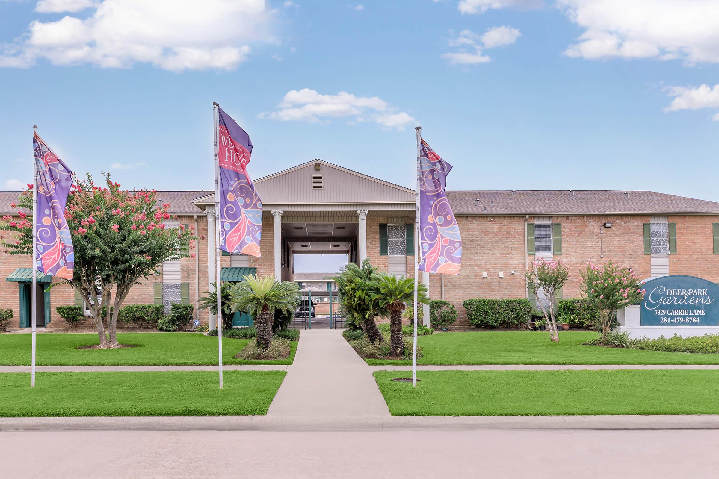 View of a residential building with neatly landscaped grounds, featuring four colorful flags in front, ornamental trees, and well-maintained grass. The entrance is marked by a spacious pathway leading to the main door, with a sign on the right displaying the name "Cedar Park Garden." The sky is partially cloudy.