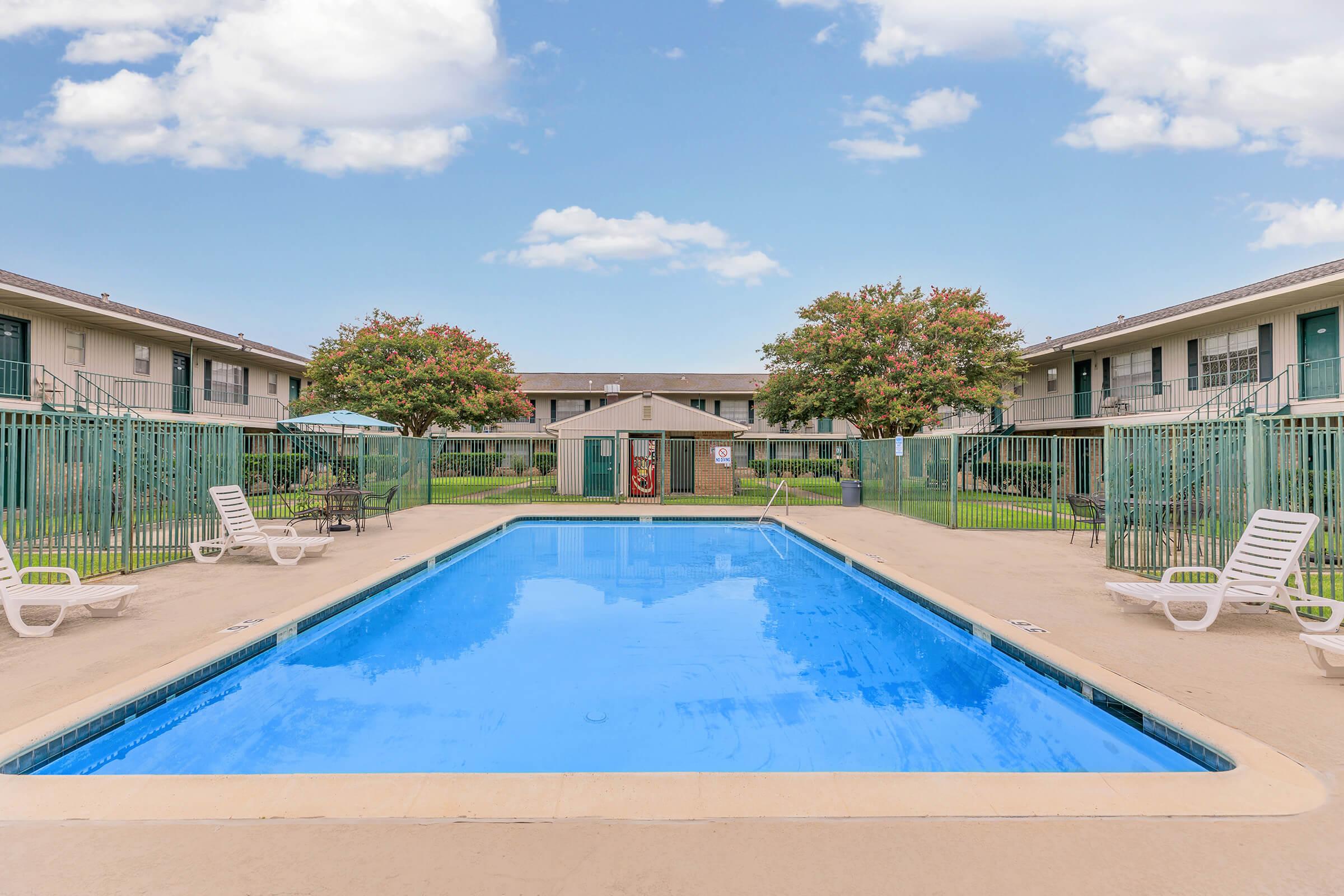 A clear blue swimming pool surrounded by lounge chairs, with a well-maintained apartment complex in the background. Trees with green foliage are visible on either side of the pool area under a bright blue sky with fluffy white clouds.