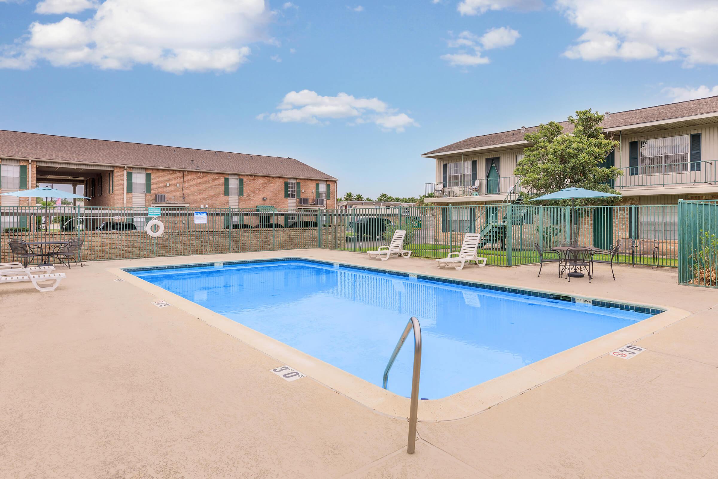 A clean outdoor swimming pool surrounded by lounge chairs and tables with umbrellas. The pool area is enclosed by a fence, and nearby buildings are visible in the background under a clear blue sky with a few clouds.