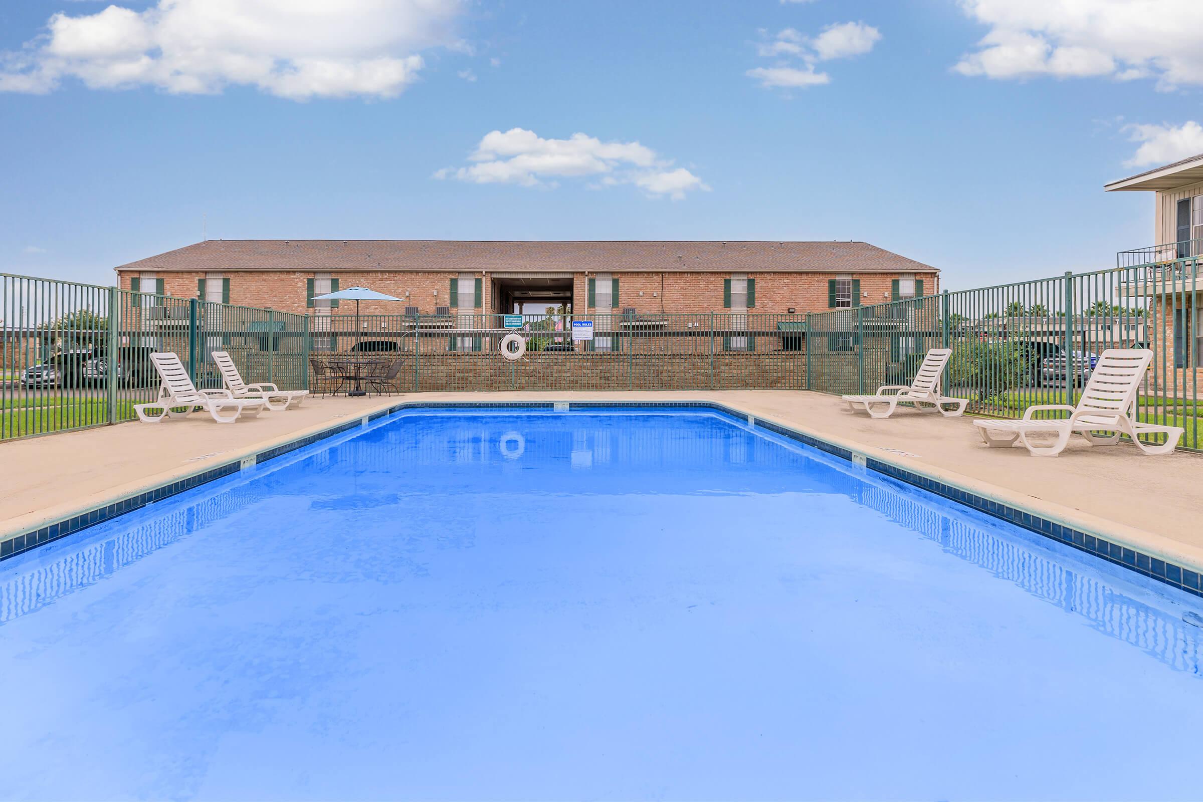 A clear blue swimming pool surrounded by lounge chairs, with a brick building in the background. Lush green grass and a few clouds in the sky create a relaxing outdoor atmosphere. The pool area is fenced for privacy and safety.