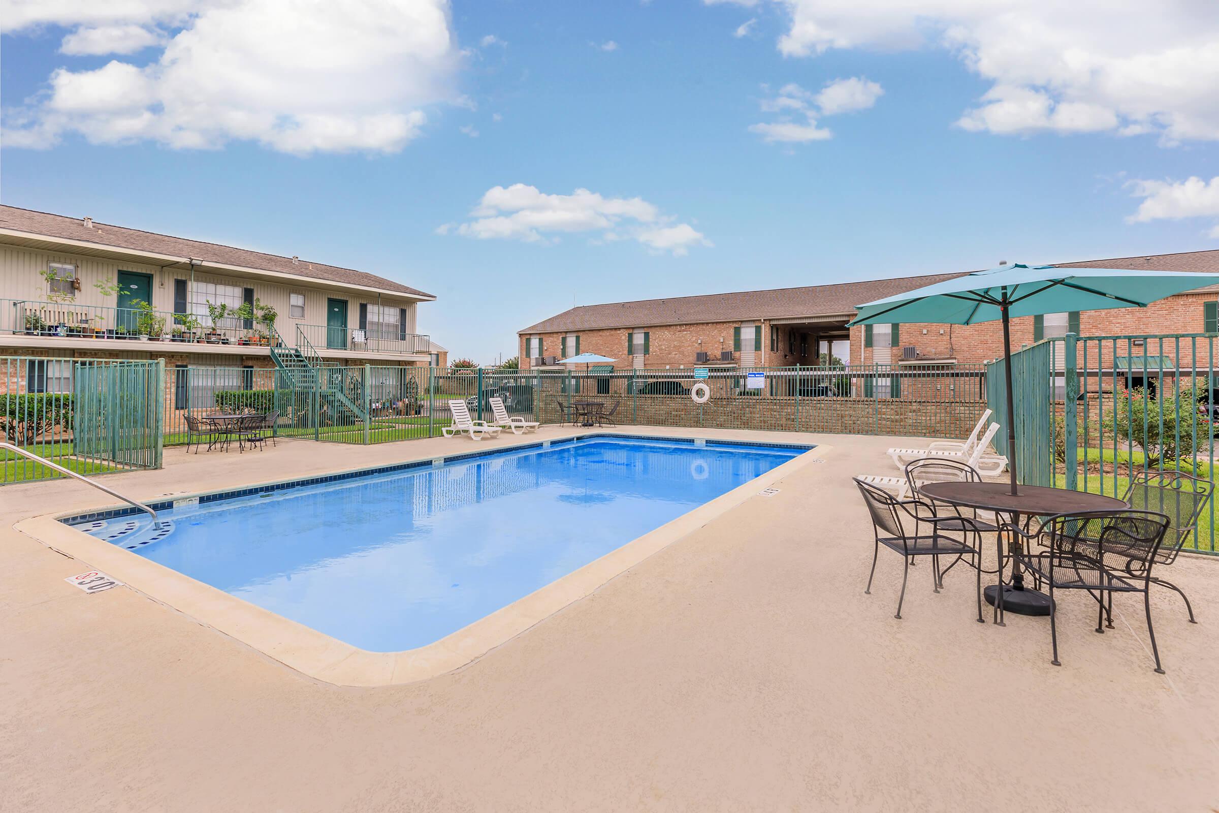 A clean, inviting outdoor swimming pool surrounded by lounge chairs and tables. In the background, there are two residential buildings with balconies. The sky is bright blue with a few fluffy clouds, creating a welcoming atmosphere.