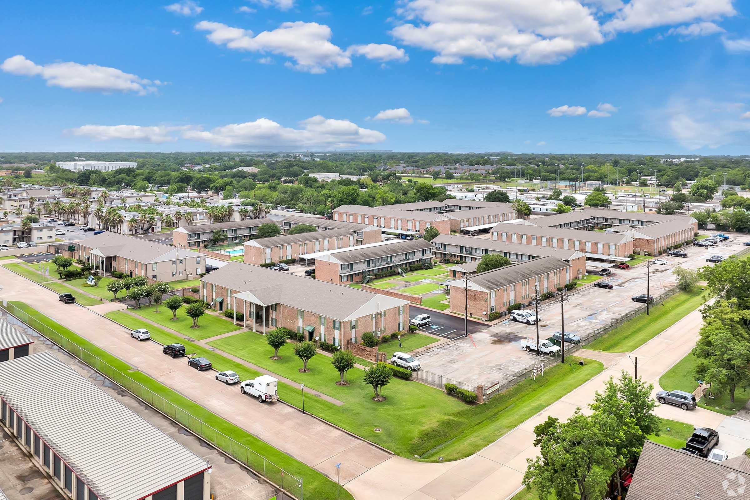 Aerial view of a residential complex featuring multiple buildings with flat roofs surrounded by green lawns and parked cars. The scene is set against a clear blue sky with a few clouds, with nearby roads and additional buildings visible in the background.