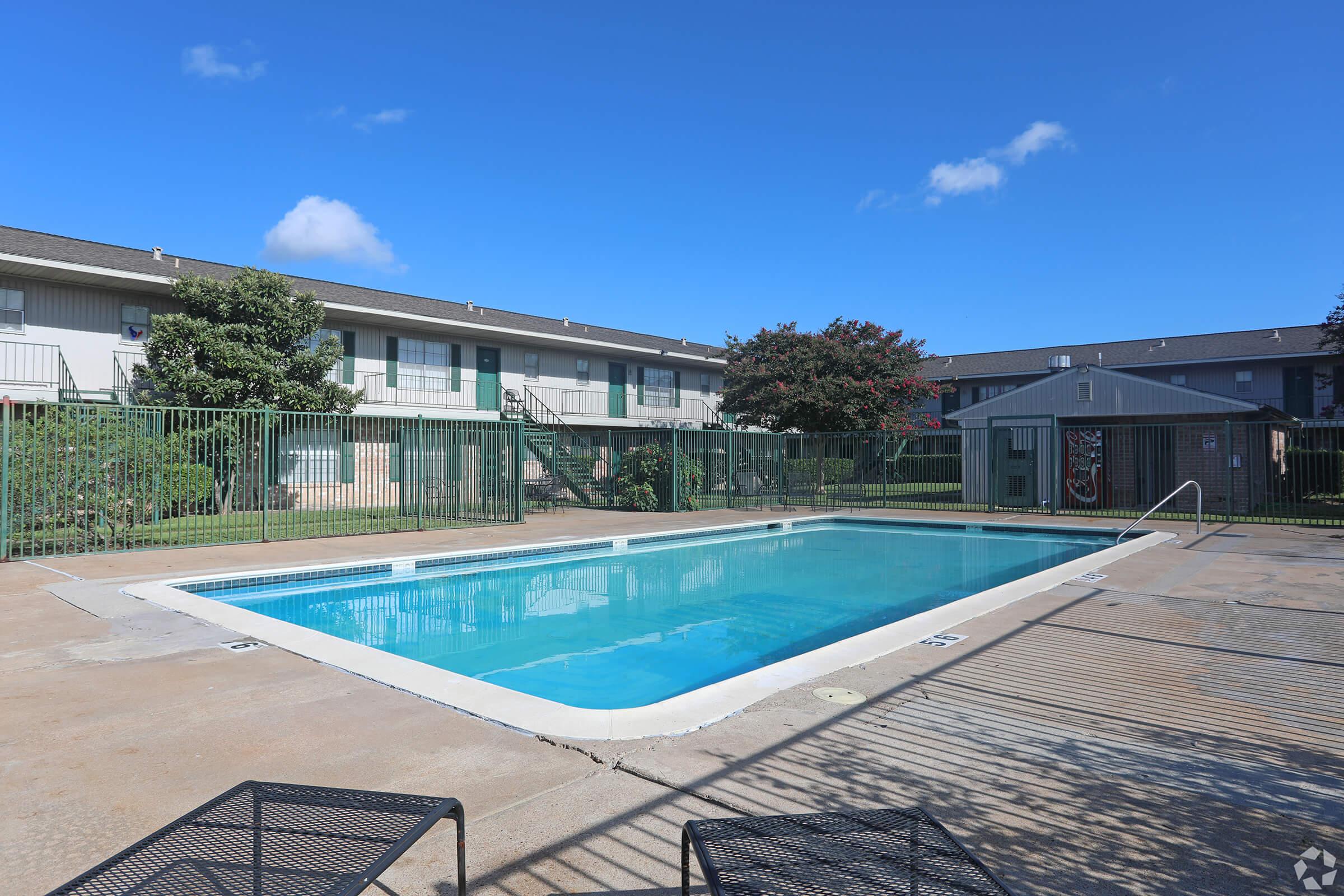 A view of an outdoor swimming pool surrounded by a fenced area, with a few poolside lounge chairs. In the background, there are two multi-story apartment buildings and some greenery, under a clear blue sky with a few clouds.