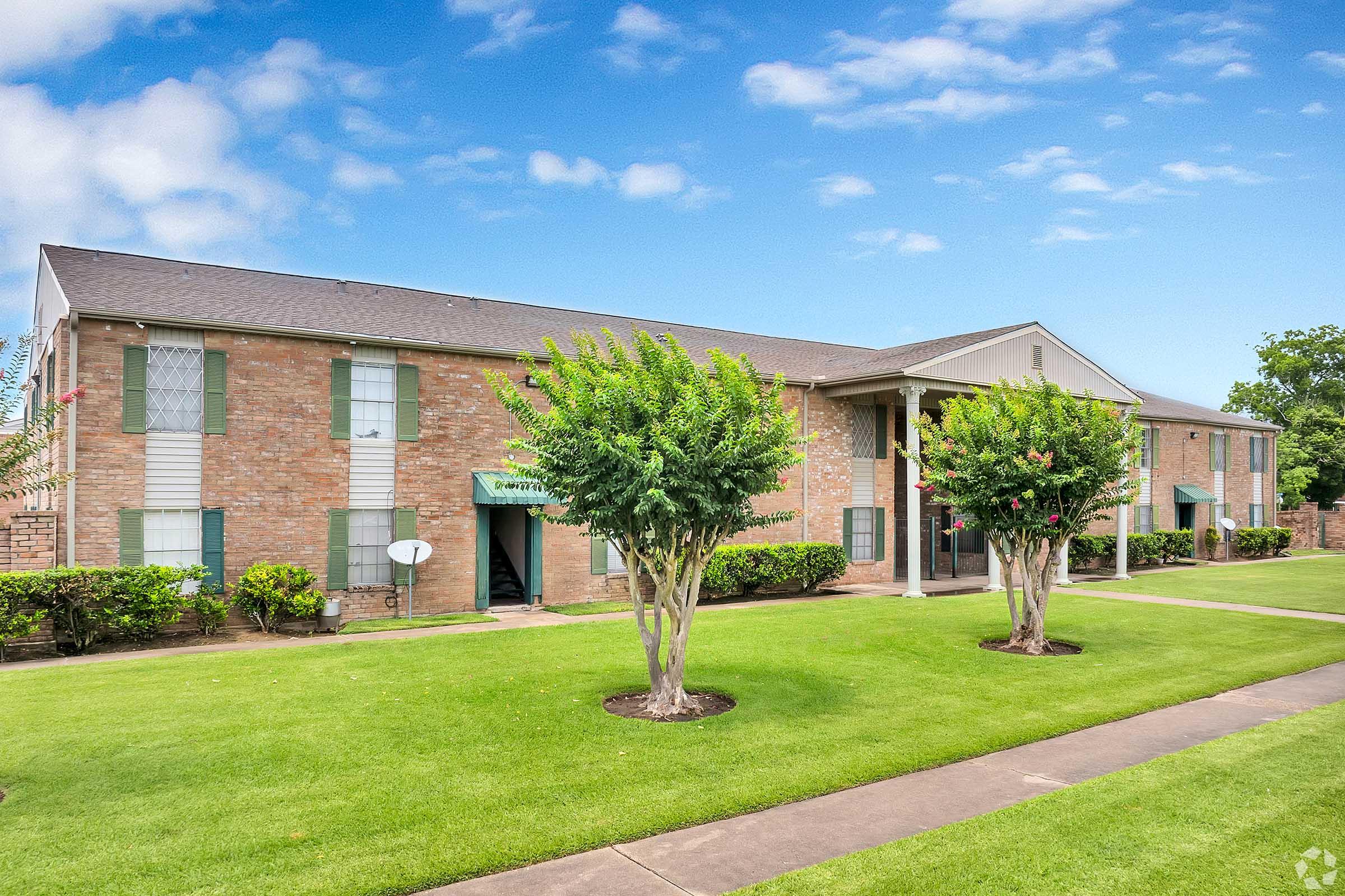 A brick apartment building with multiple units featuring green shutters, surrounded by well-maintained lawns and small trees. The sky is clear with a few clouds, and a sidewalk runs in front of the building, enhancing the residential setting.