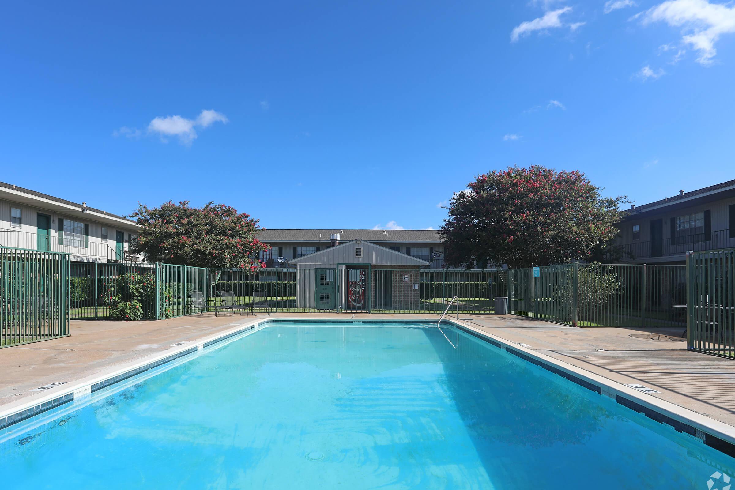 A clear outdoor swimming pool surrounded by a green fence, with a view of two multi-story apartment buildings in the background. Bright blue sky with a few clouds contrasts against the serene setting. Lush green trees line the pool area, creating a peaceful atmosphere.