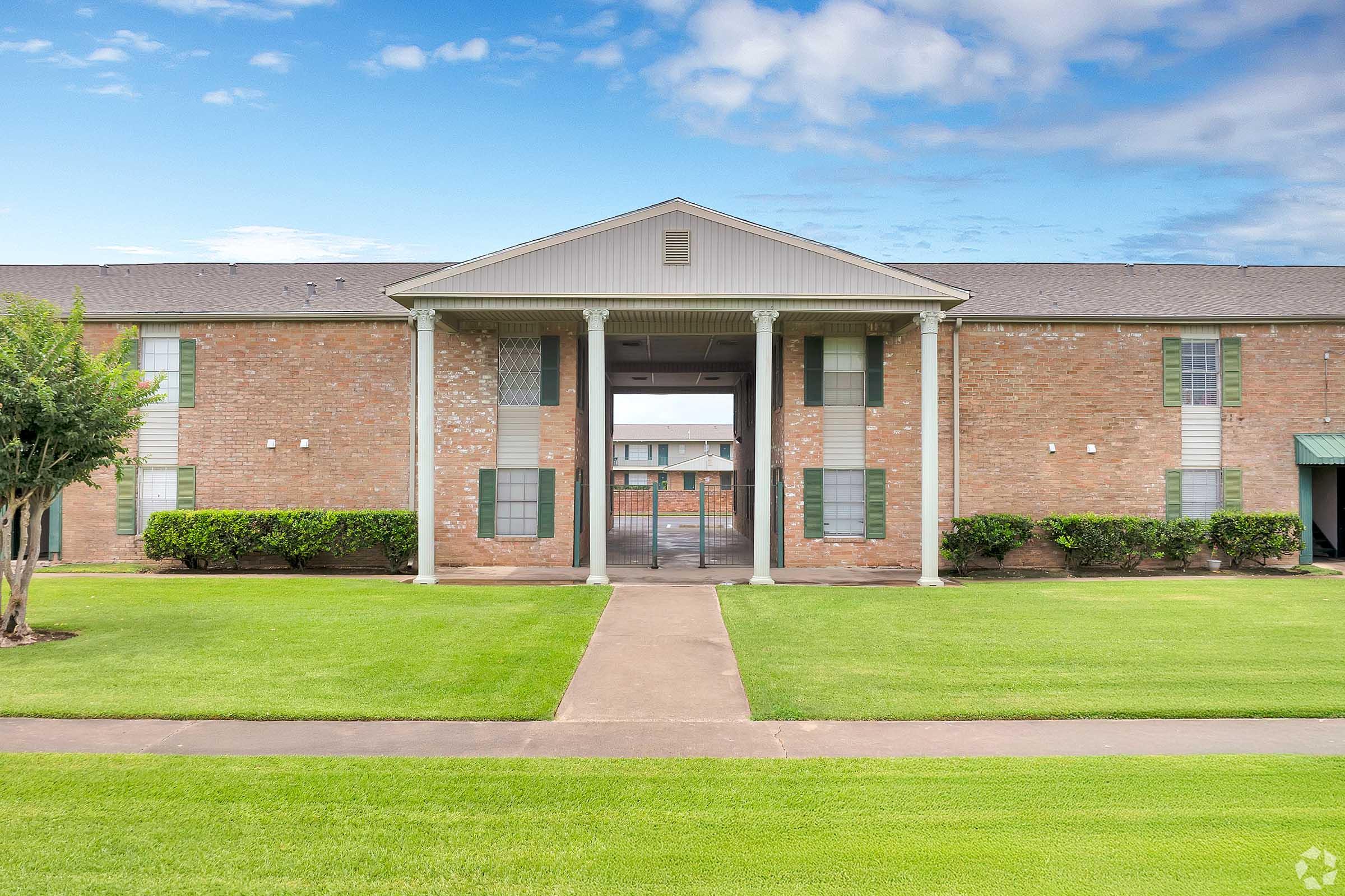 Exterior view of a two-story residential building with brick walls and green accents. The entrance features a covered porch with white columns. Well-maintained lawn and pathway leading to the entrance. The sky is partly cloudy, creating a bright atmosphere.