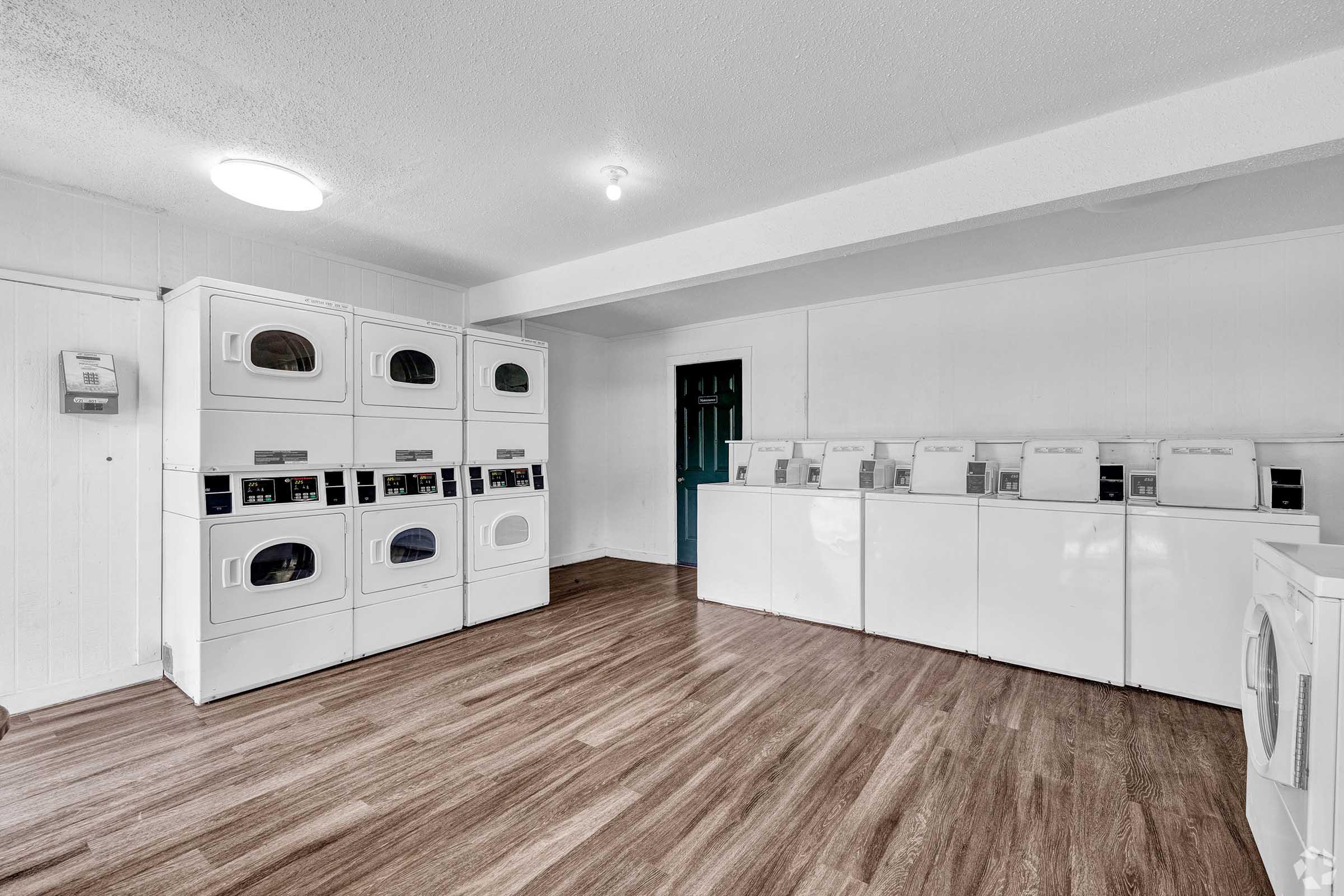 A clean, spacious laundry room featuring multiple white washing machines and dryers lined against the walls. The floor is wood-like, and there's a door leading to another room. Adequate lighting enhances the bright and organized atmosphere.