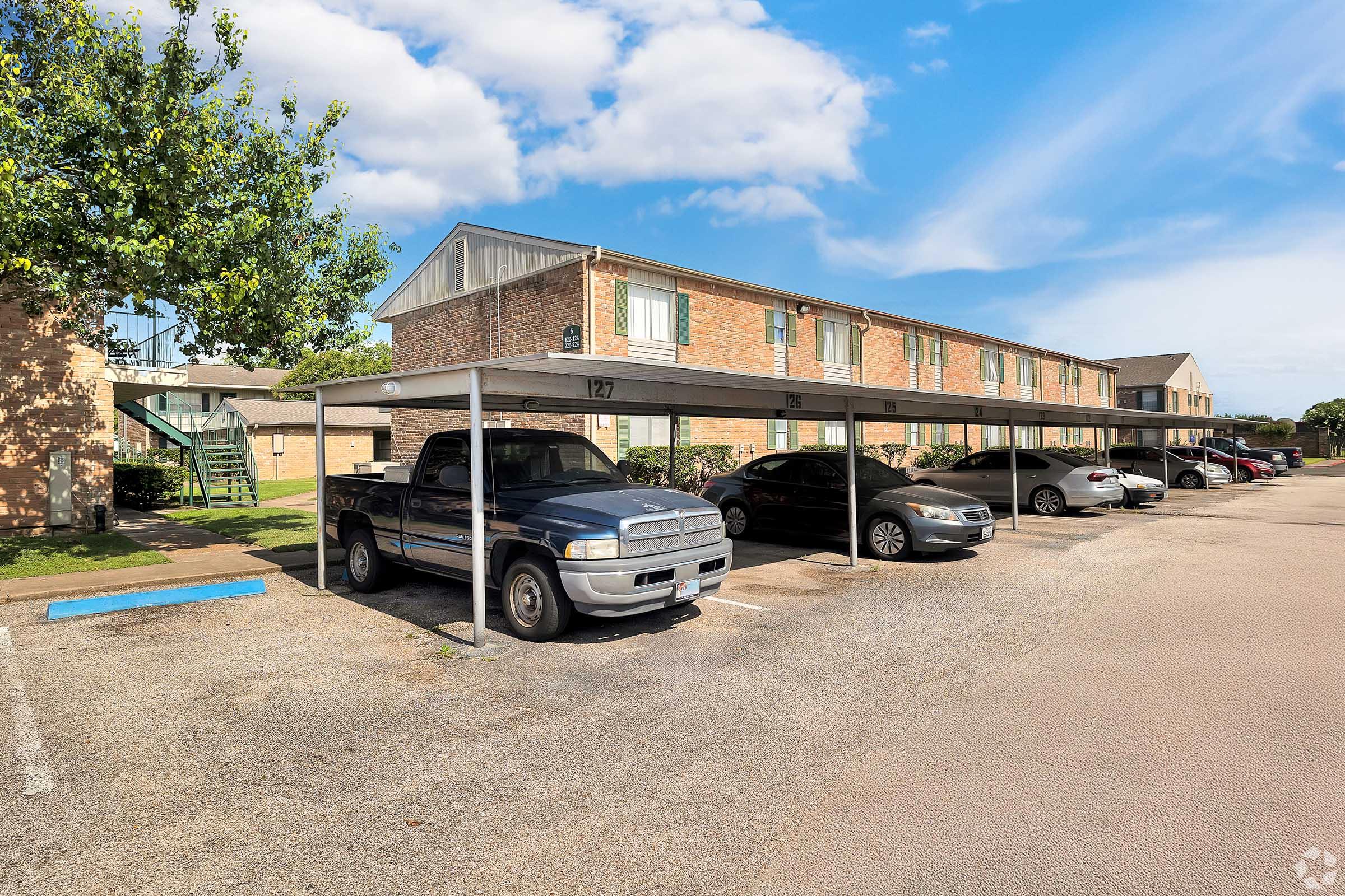 A parking area with several covered spaces. A silver pickup truck is parked in the foreground, along with a few other vehicles. In the background, there are brick apartment buildings with green balconies and trees nearby under a clear blue sky.