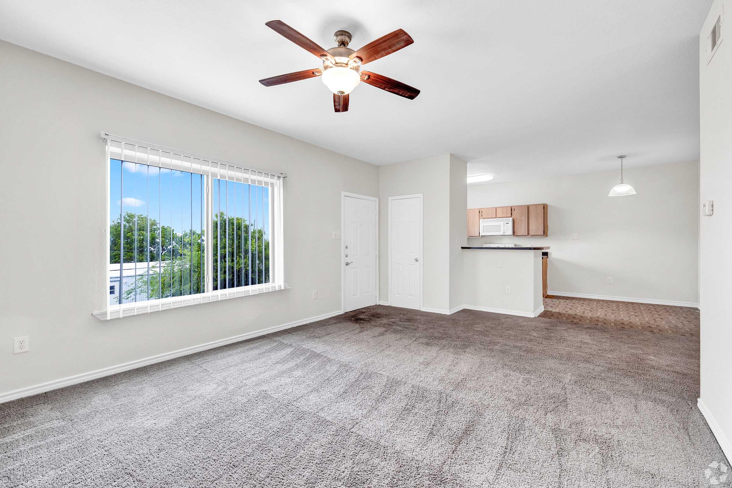 Spacious living area with beige carpet, ceiling fan, and large window allowing natural light. White walls and a doorway leading to a kitchen with wooden cabinets are visible in the background. The overall vibe is bright and airy, perfect for comfortable living.