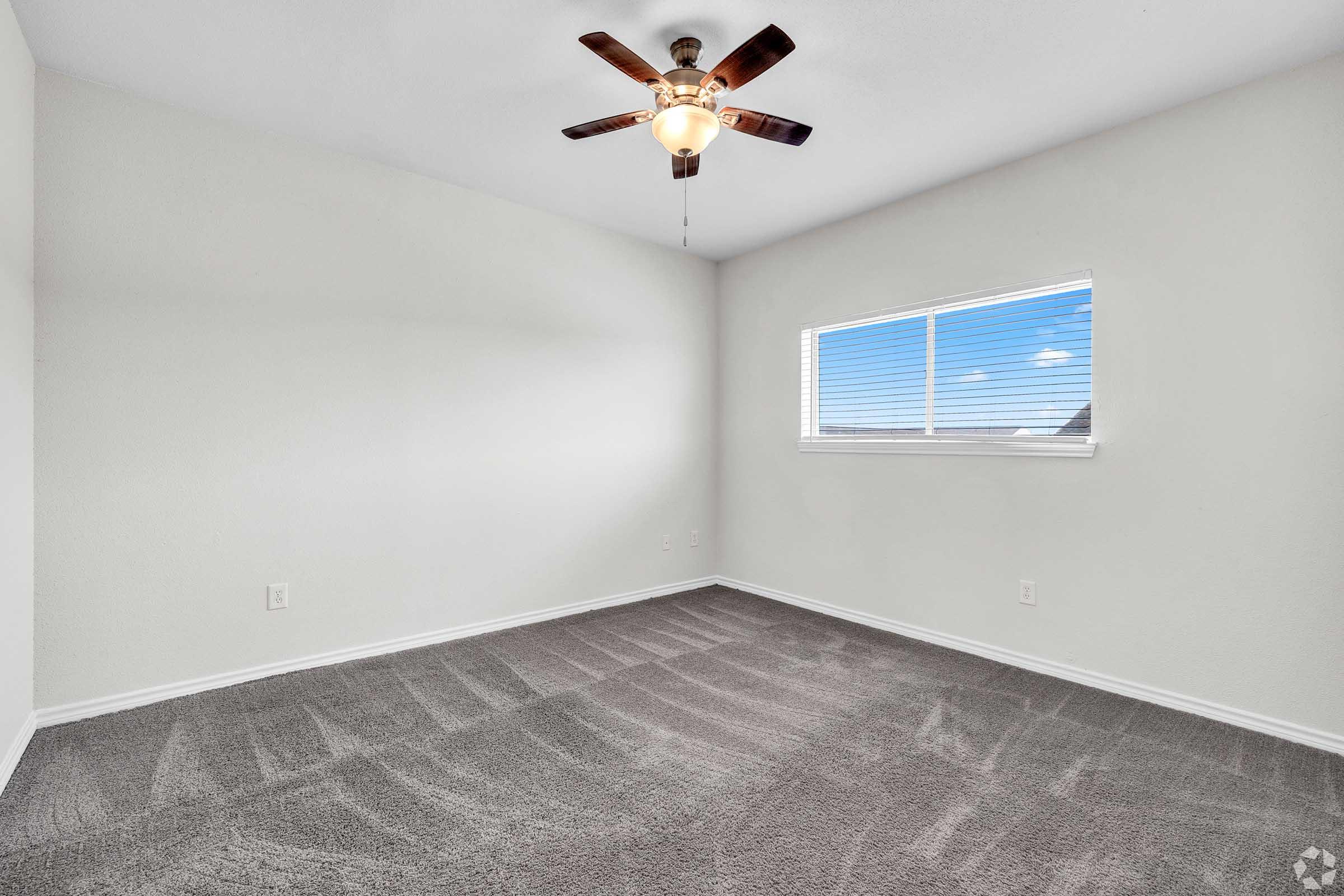 A spacious, empty room with light gray carpet and freshly painted white walls. A ceiling fan is installed, and a window with white blinds allows natural light to fill the space. The room is well-lit and airy, offering a clean and neutral environment, ideal for various uses or personal customization.
