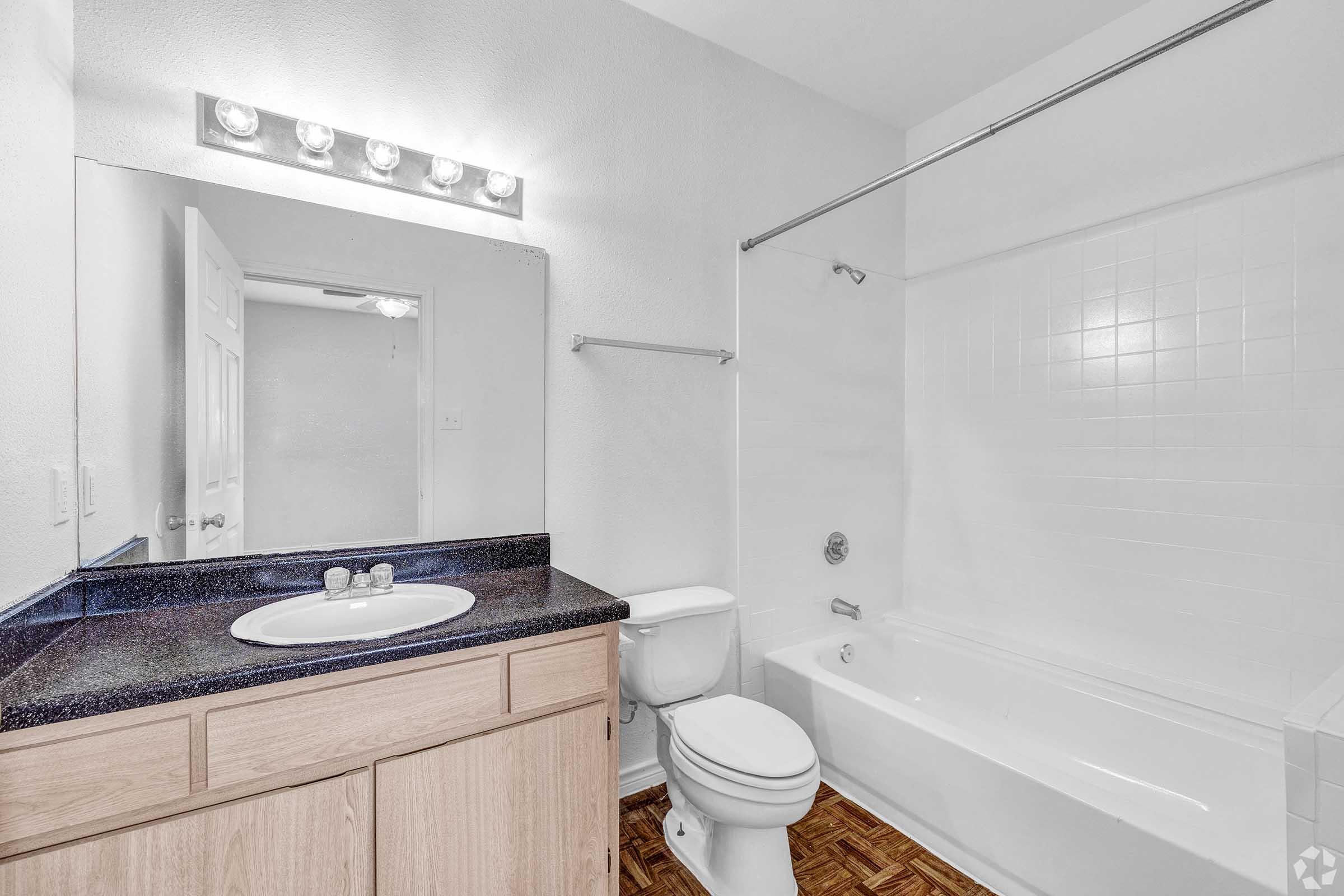A clean and spacious bathroom featuring a mirrored vanity with a single sink, wooden cabinetry, a toilet, and a bathtub with a shower. The walls are painted white, and the floor has a wood-like texture. Bright lighting above the mirror enhances the overall brightness of the room.