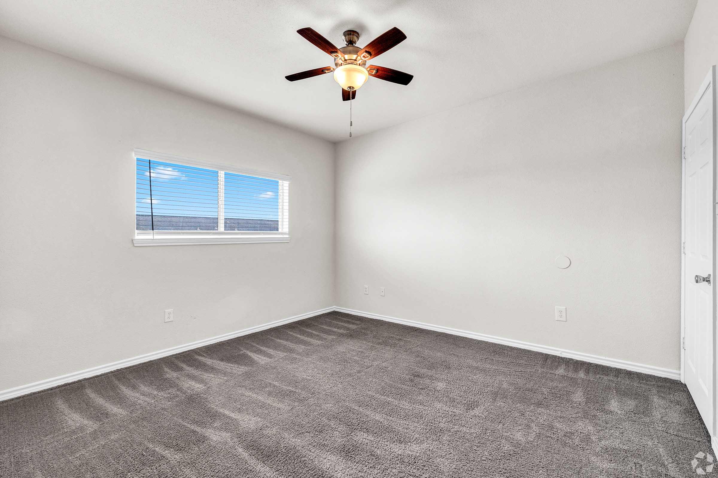 Empty room with light gray carpet and white walls. A ceiling fan with wooden blades is installed in the center. There is a window with blinds allowing natural light to enter, illuminating the space. The room appears ready for furniture placement and personalization.