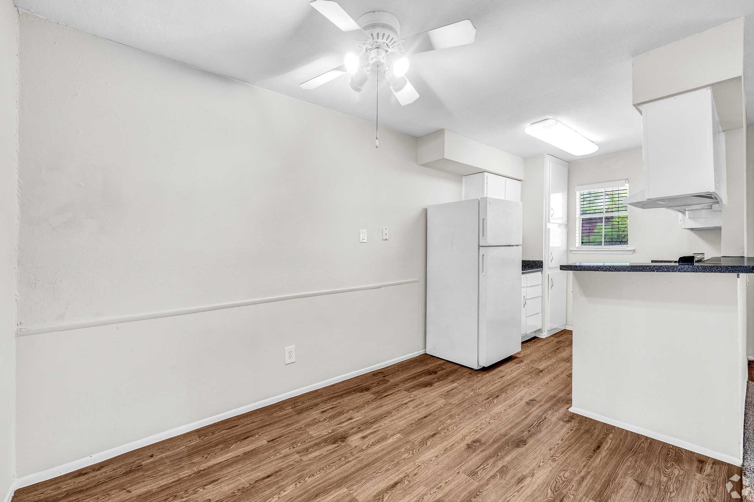 A well-lit kitchen area featuring a white refrigerator, light wood flooring, and light-colored walls. A ceiling fan is visible, and there is an open layout with a countertop and window letting in natural light. The space appears clean and inviting, suitable for casual dining or cooking.