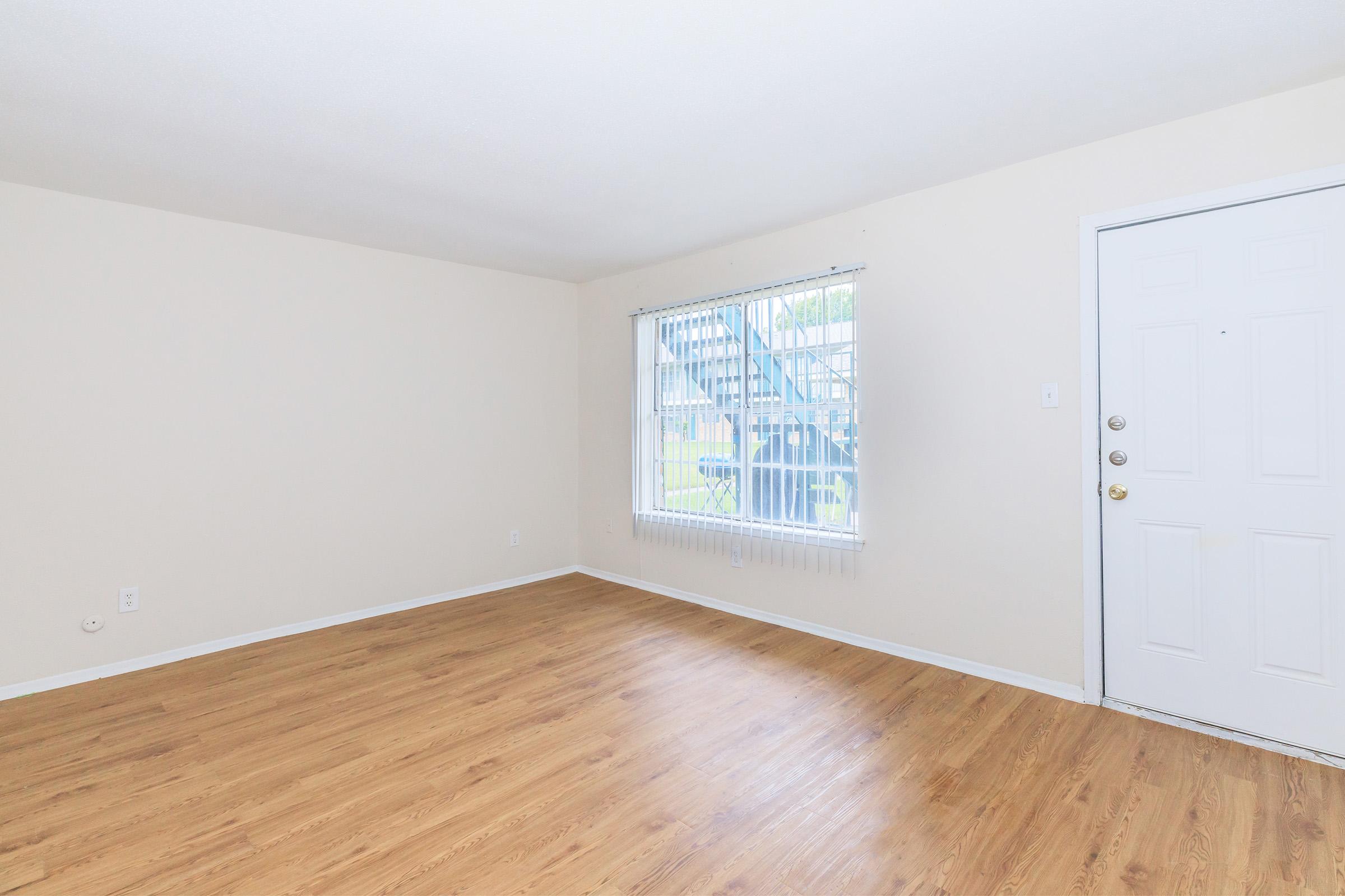 A spacious, empty living room with light-colored walls and wooden flooring. A large window lets in natural light, offering a view of a staircase outside. The room features a front door with multiple locks and a simple light fixture on the ceiling, creating a bright and welcoming atmosphere.