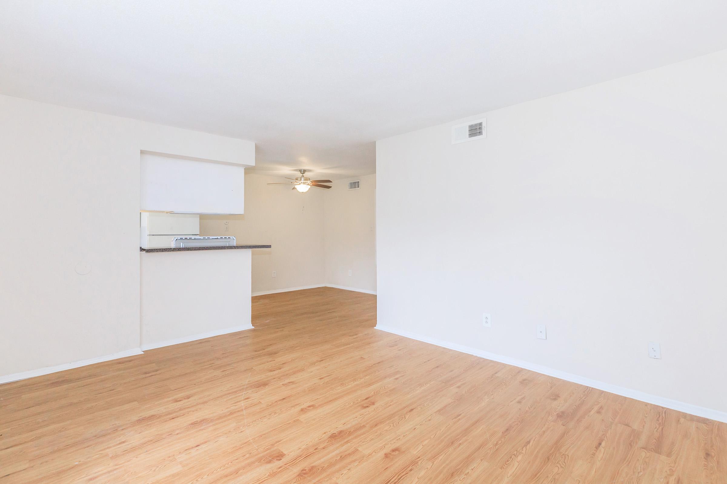 Spacious, empty living area featuring light-colored walls and a wood-style floor. A ceiling fan is visible, and there is an open kitchen area in the background with a countertop. Natural light fills the space, creating a bright and inviting atmosphere.