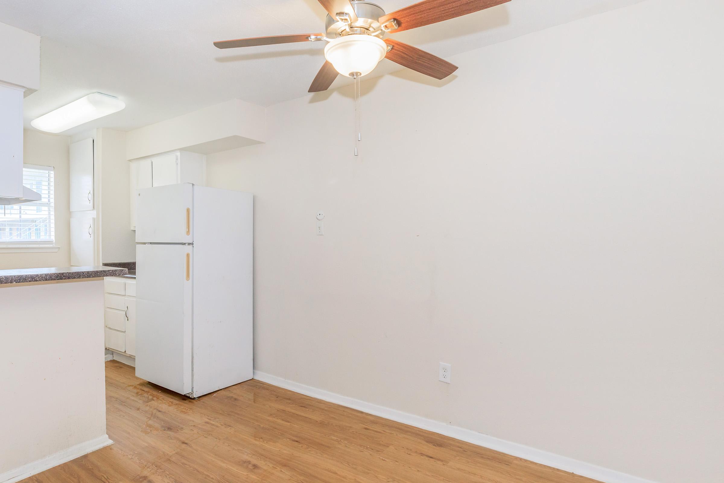 A small kitchen area featuring a white refrigerator, light-colored cabinets, and a light wood floor. A ceiling fan is visible, and the walls are painted a soft neutral color. Natural light enters through a nearby window, creating a bright and open atmosphere.