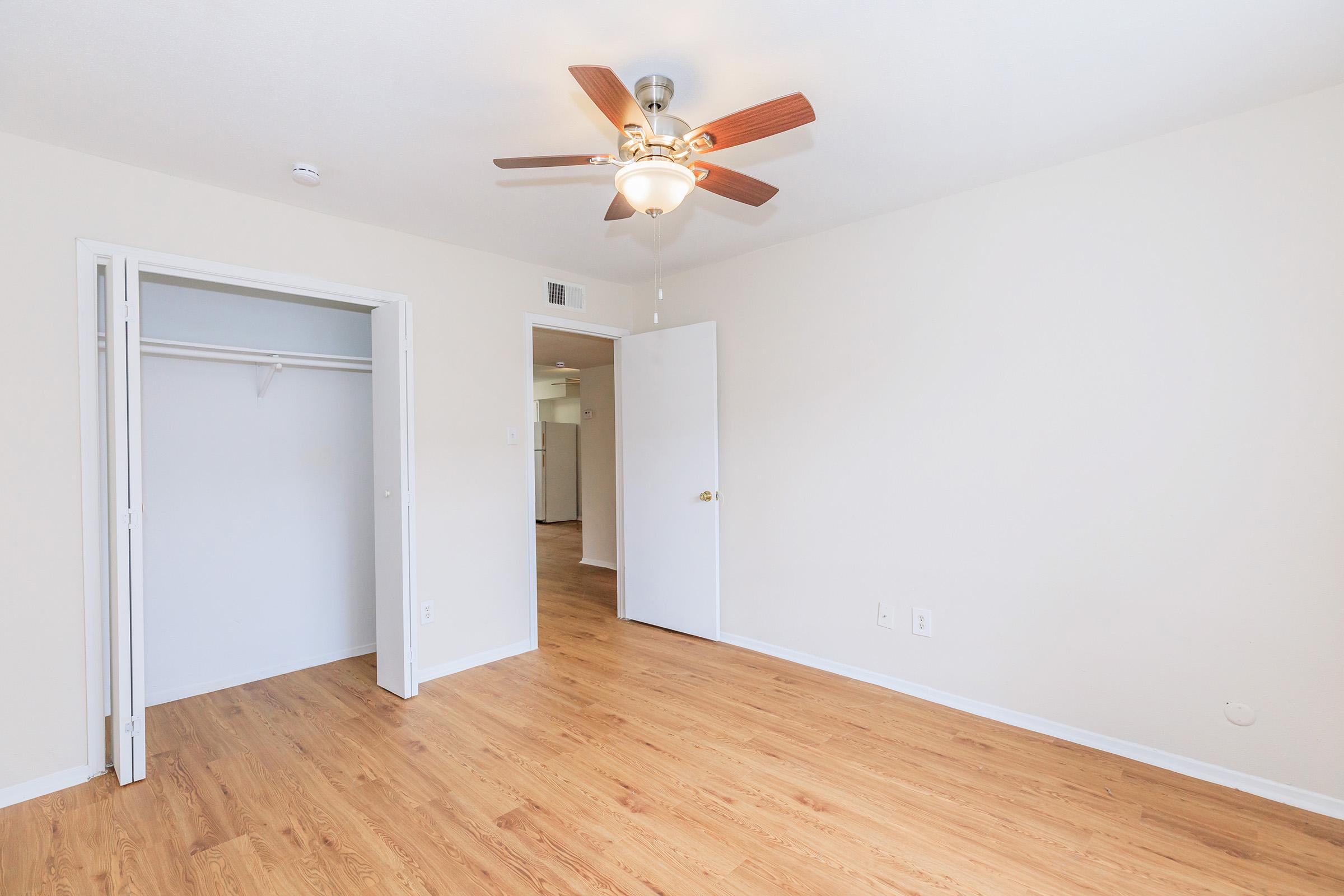 A light-colored room with wooden flooring, featuring a ceiling fan. There is an open closet to the left and a closed door leading to another area. The walls are painted a neutral shade, creating a bright and spacious atmosphere.